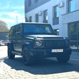 My Clean. Car Premium Detailing - Czarny, matowy Mercedes-Benz G-Class zaparkowany na brukowanym placu, z widocznym budynkiem w tle i fragmentem stadionu po lewej stronie. Tablica rejestracyjna z polską flagą.