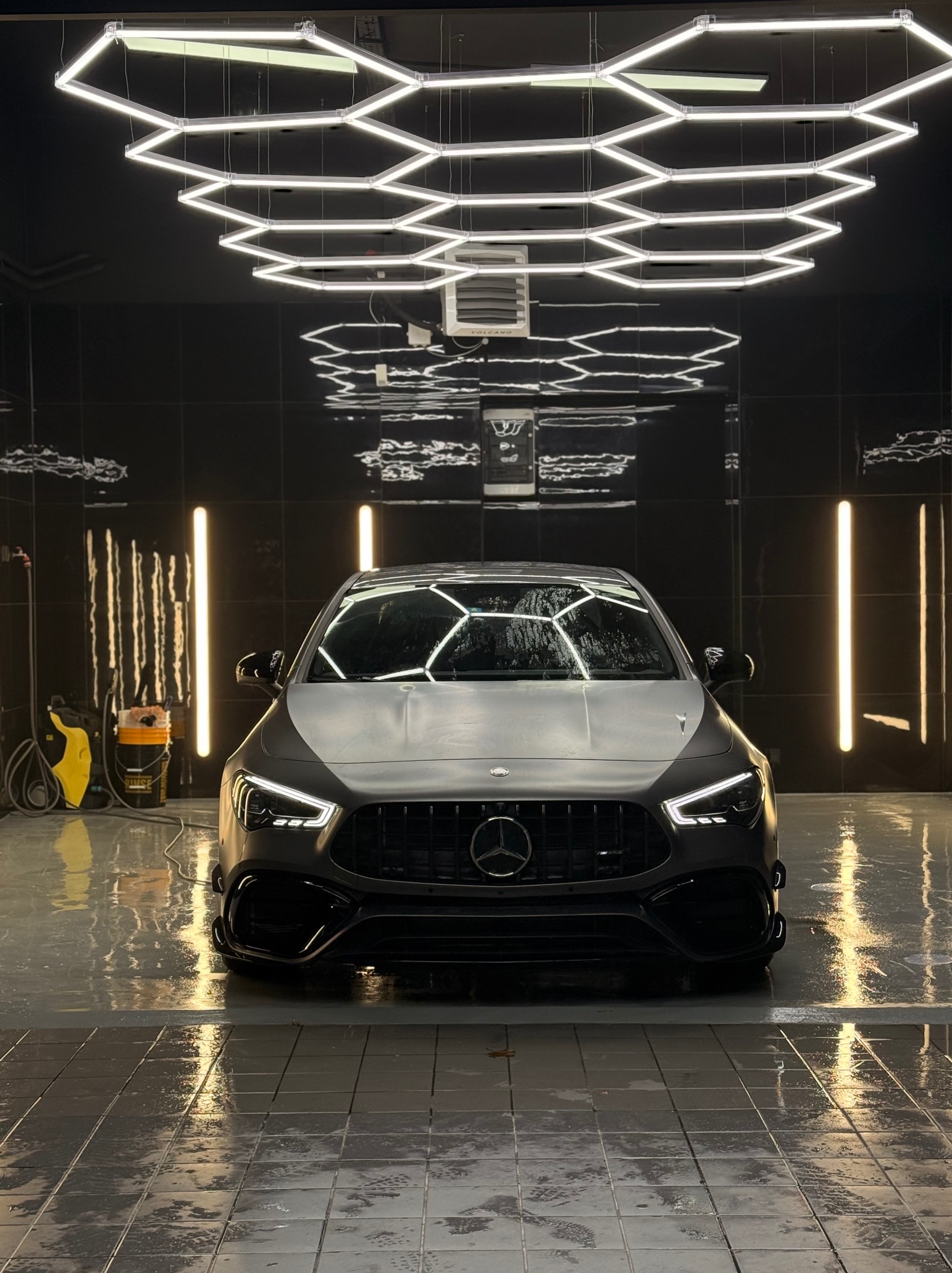 Czarny Mercedes-AMG w myjni z heksagonalnym oświetleniem sufitowym i mokrą posadzką, odbijającą światła. Widok z przodu, detale grilla i reflektorów.