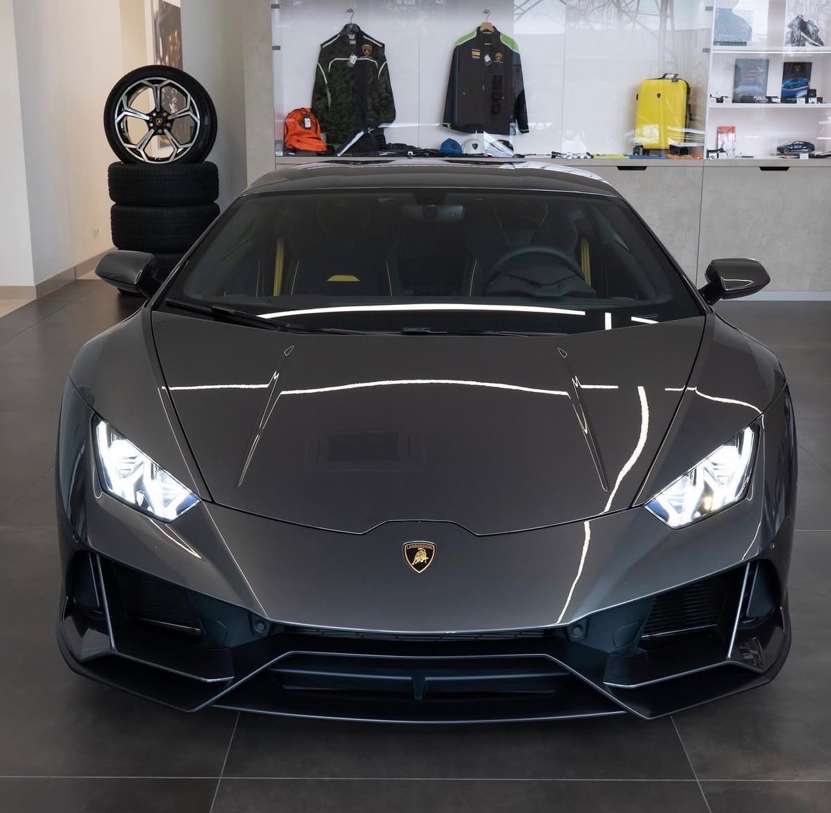 Szary Lamborghini Huracan z włączonymi światłami, widok z przodu, na tle opon i ubrań na wystawie w salonie. Widoczne logo Lamborghini.