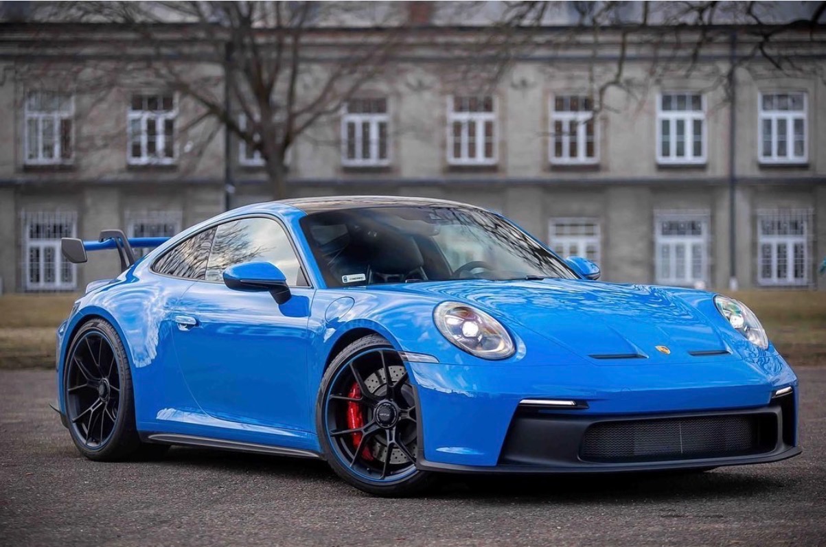 Błękitne Porsche 911 GT3 na tle budynku z szarymi ścianami i białymi oknami. Widoczne czarne felgi z czerwonymi zaciskami hamulcowymi. Auto po detailingu.
