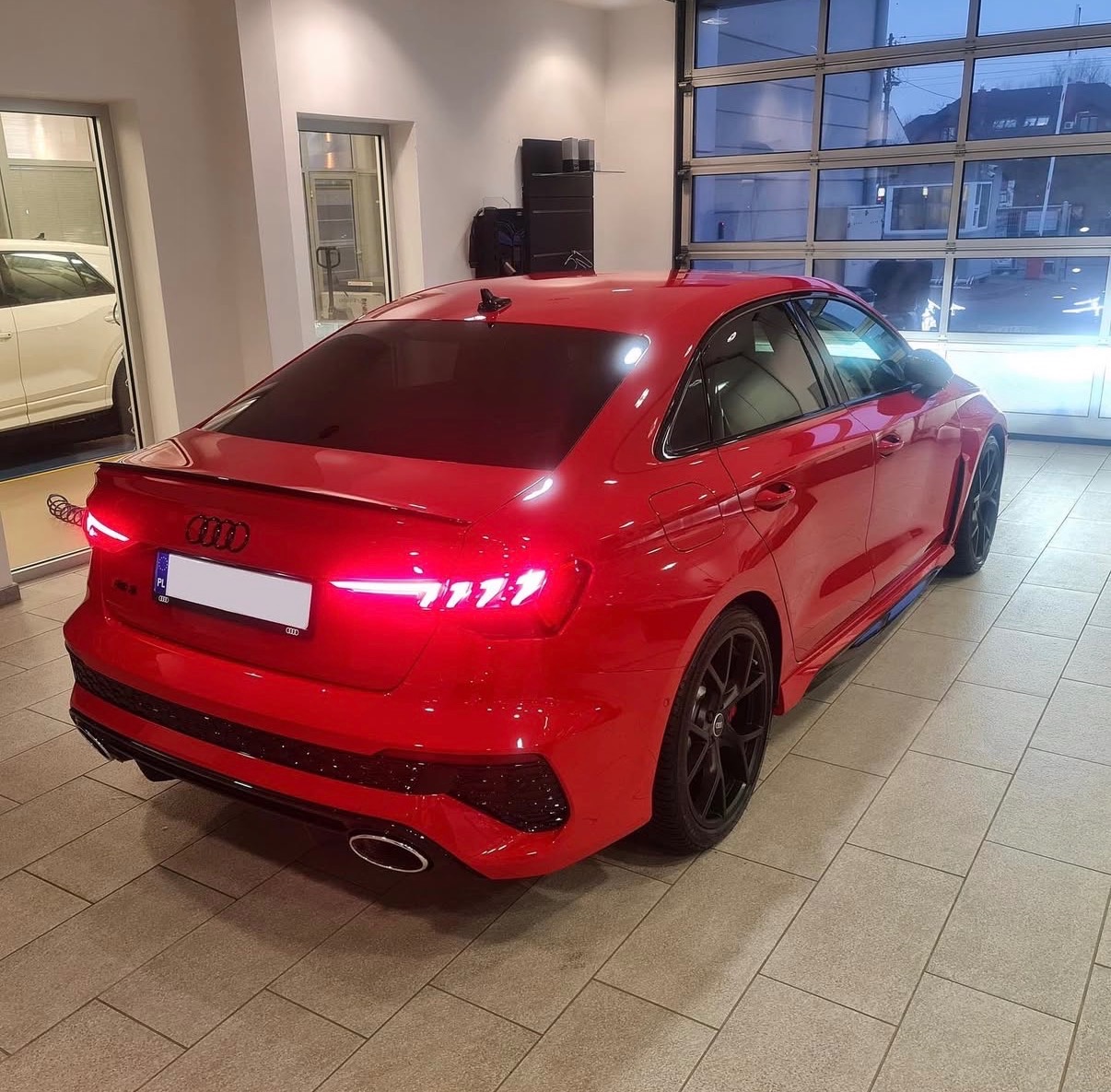 Czerwone Audi RS3 po oklejeniu folią, widoczne czarne felgi i przyciemnione szyby. Auto stoi w jasnym pomieszczeniu z przeszkloną ścianą.