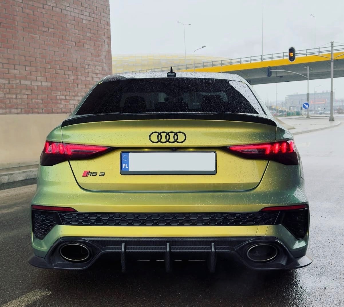 Żółto-zielony Audi RS3 po oklejeniu folią, widok z tyłu na mokrej nawierzchni. Widoczne detale: czarny dyfuzor, logo Audi i oznaczenie modelu.