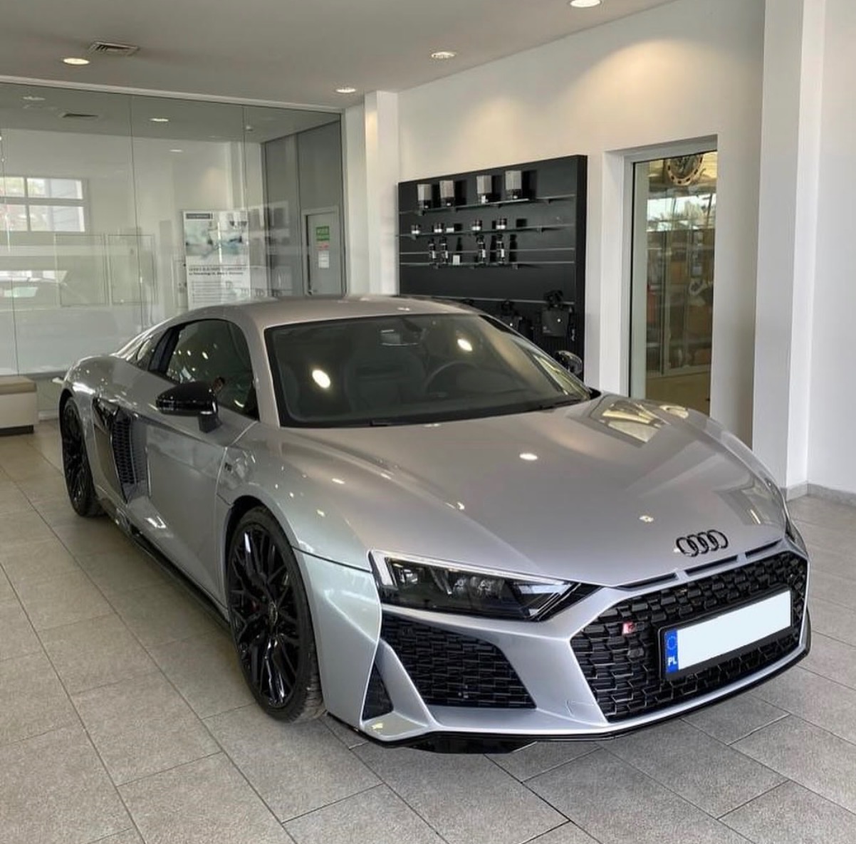 Srebrne Audi R8 na tle jasnego salonu z przeszklonymi ścianami i czarnymi półkami. Sportowy samochód z czarnymi felgami prezentuje się elegancko.