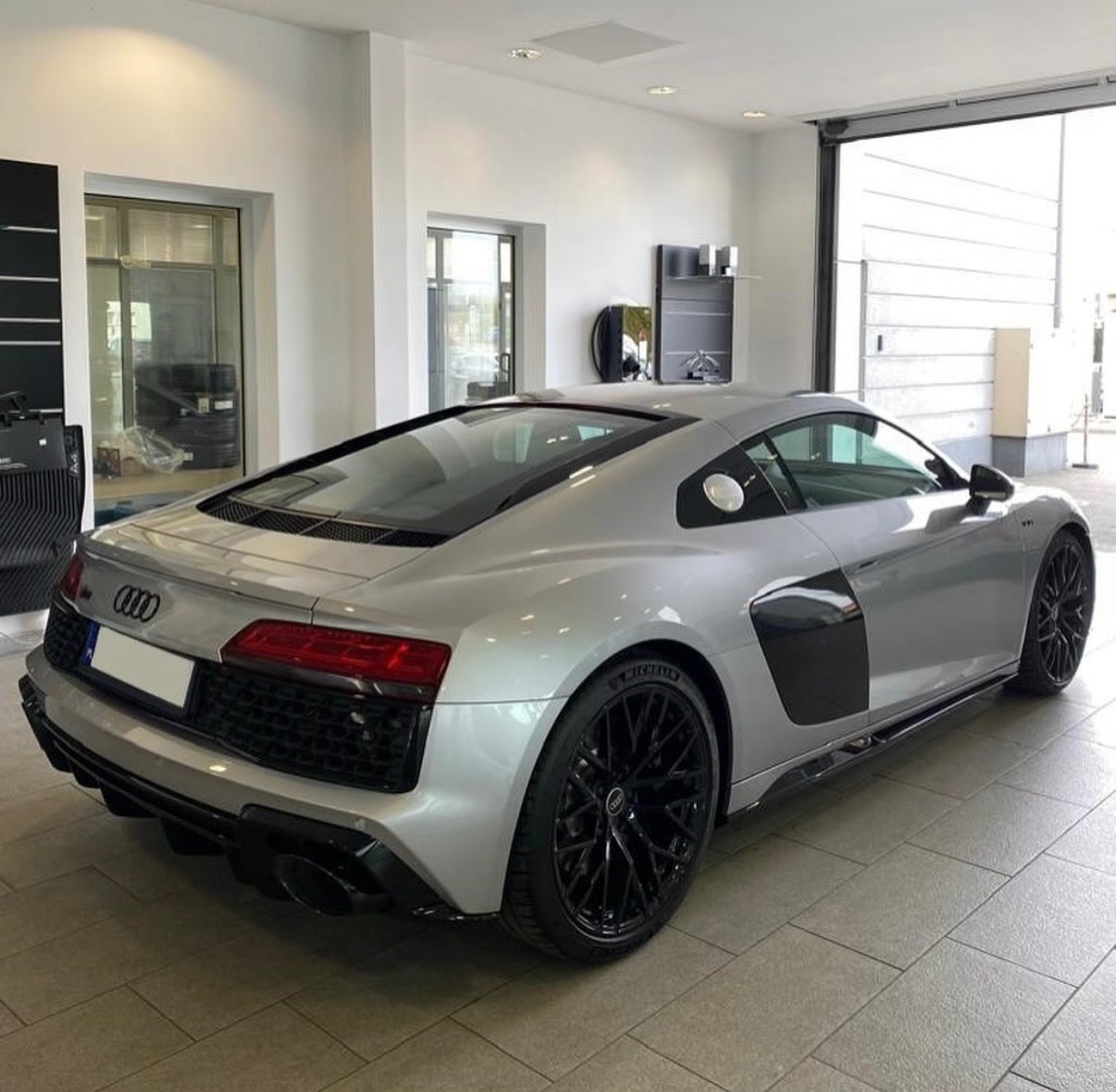 Srebrne Audi R8 z czarnymi felgami, widziane z tyłu, w jasnym salonie. Widoczne detale karoserii i oświetlenia.