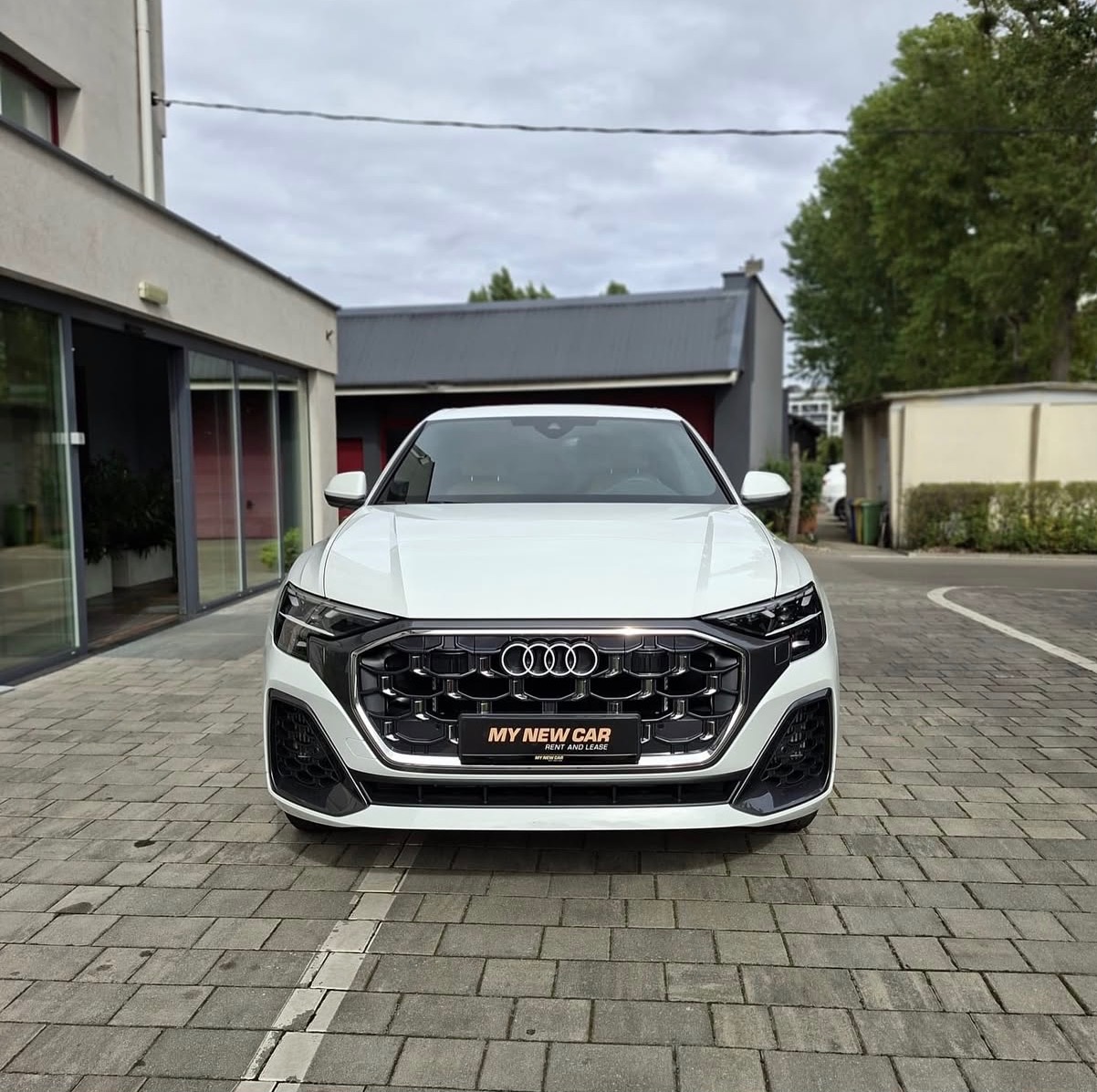 Białe Audi Q8 z widocznym logo firmy 'My New Car' na grillu, zaparkowane na brukowanej nawierzchni przed budynkiem z przeszklonymi drzwiami.