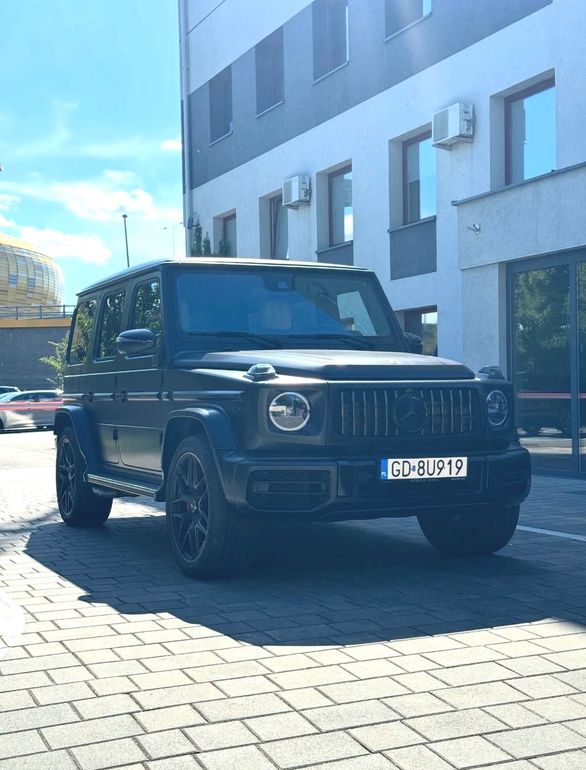 Czarny, matowy Mercedes-Benz G-Class zaparkowany na brukowanym placu, z widocznym budynkiem w tle i fragmentem stadionu po lewej stronie. Tablica rejestracyjna z polską flagą.
