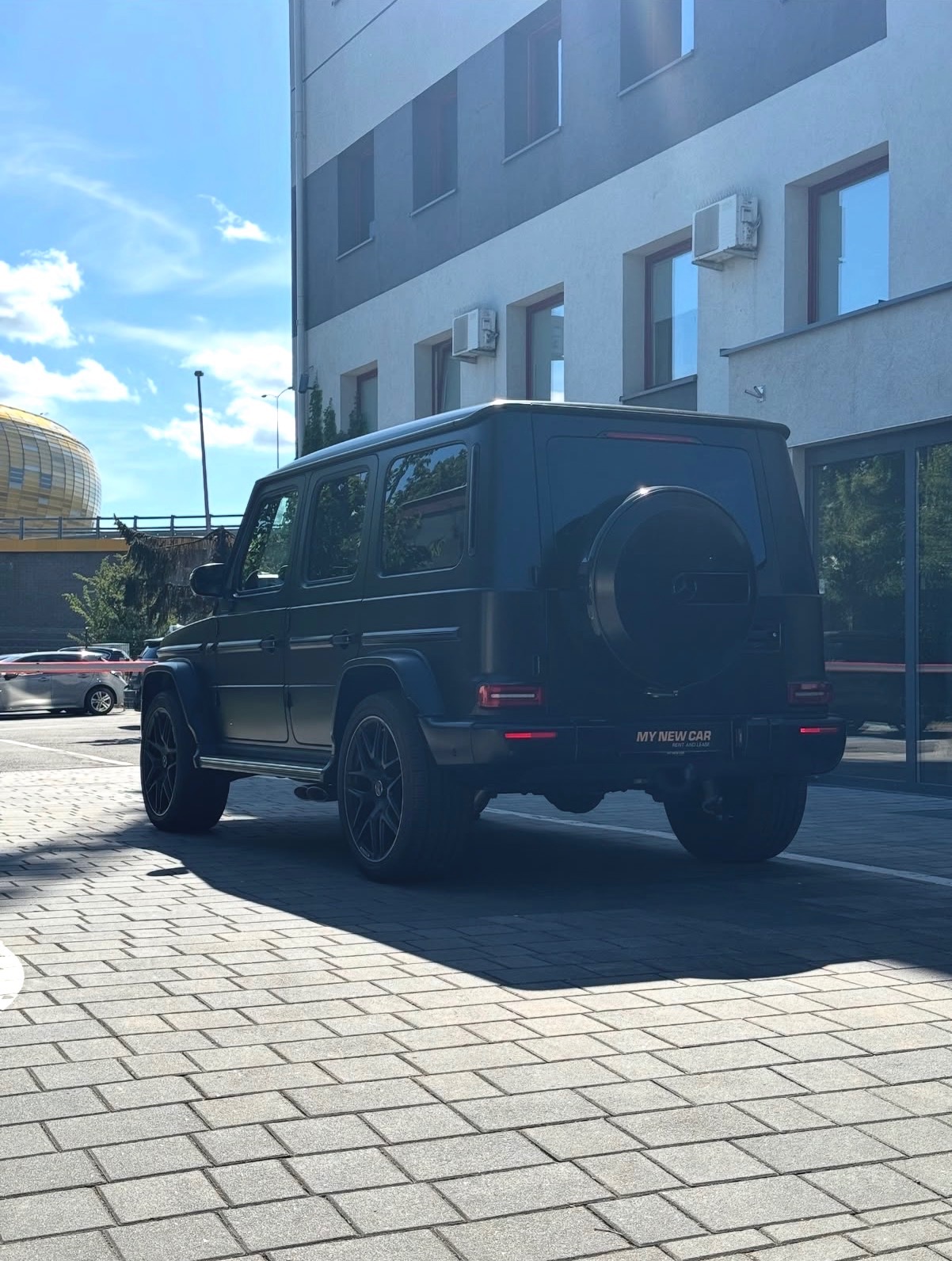 Czarny SUV z kołem zapasowym na klapie bagażnika, zaparkowany na brukowanej nawierzchni, z budynkiem w tle i widocznym fragmentem stadionu.