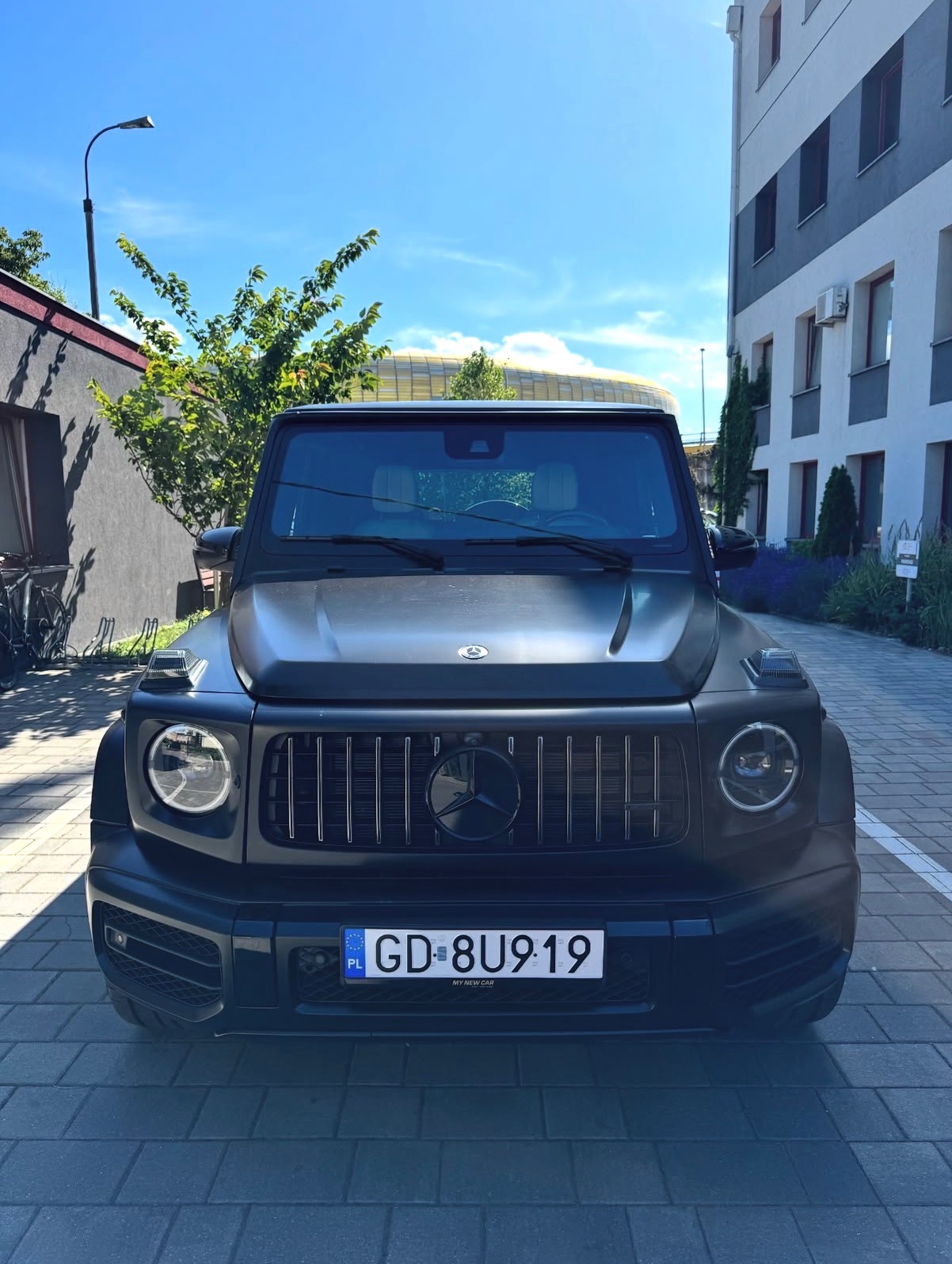 Czarny, matowy Mercedes-Benz G-Klasy zaparkowany na brukowanej drodze, z widocznym budynkiem w tle i fragmentem drzewa po lewej stronie. Tablica rejestracyjna widoczna.