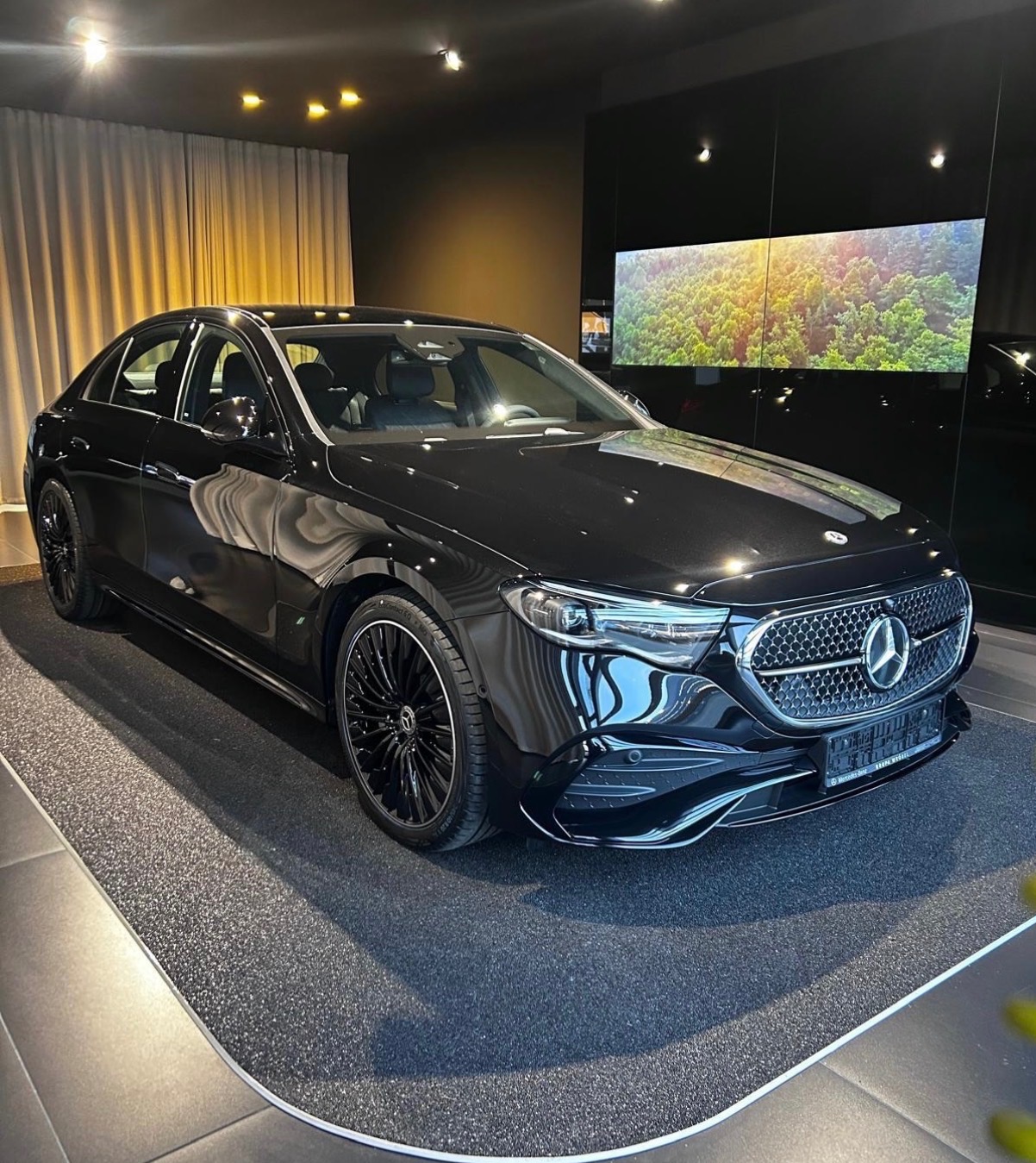 Czarny Mercedes-Benz E-Klasa z chromowanymi akcentami, prezentowany w salonie samochodowym z oświetleniem punktowym i ekranem LED z motywem leśnym w tle.