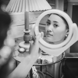 fotolodz - Czarno-białe zdjęcie kobiety przeglądającej się w okrągłym lusterku, z widoczną lampką i elementami wystroju wnętrza w tle.