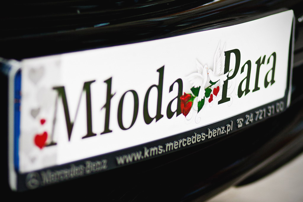 Tablica rejestracyjna z napisem 'Młoda Para' ozdobiona gołębiami, różą i sercami na czarnym samochodzie Mercedes-Benz.