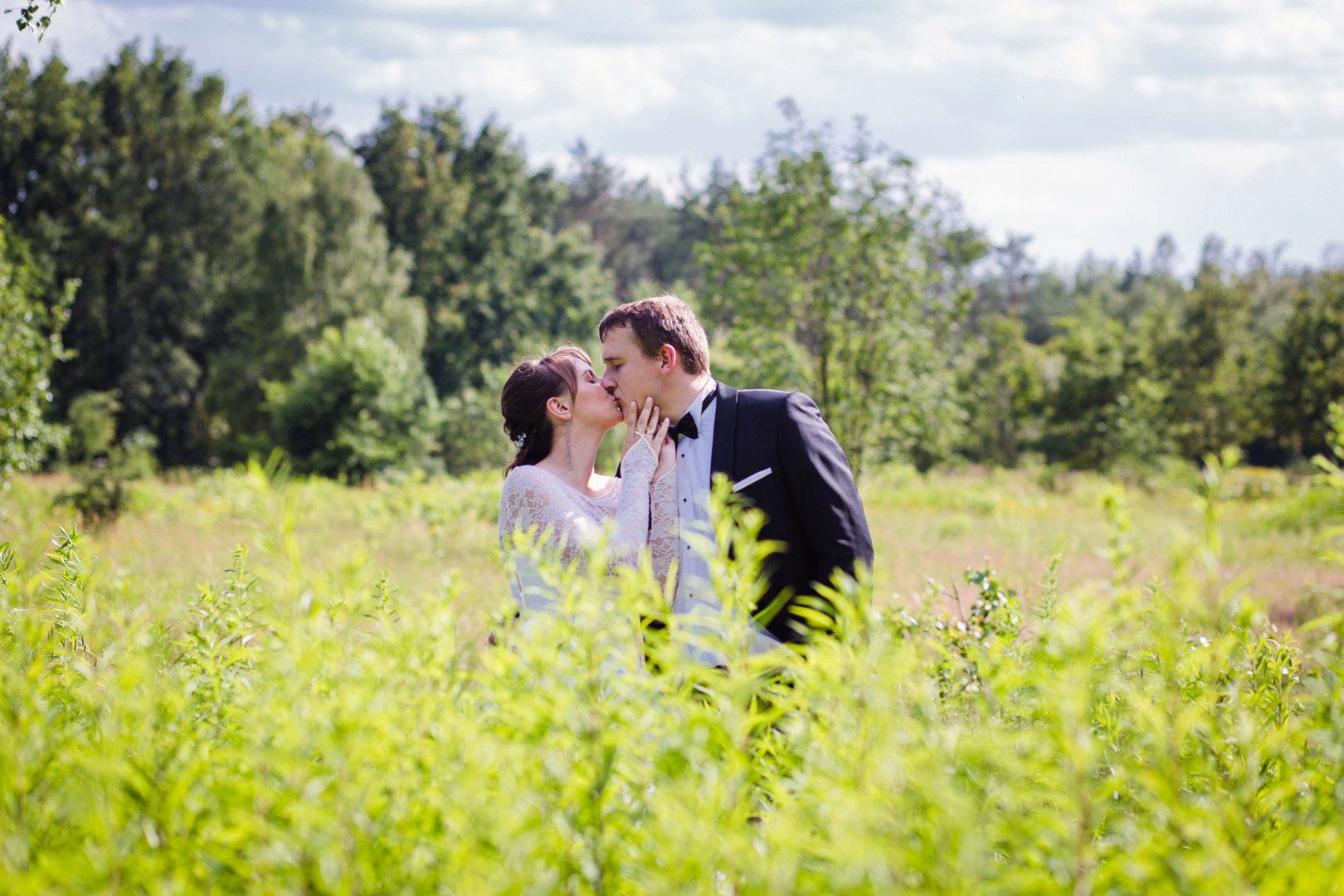Pocałunek pary młodej na tle zieleni. Kobieta w koronkowej sukni, mężczyzna w czarnym smokingu. Naturalne światło i plenerowa sceneria.