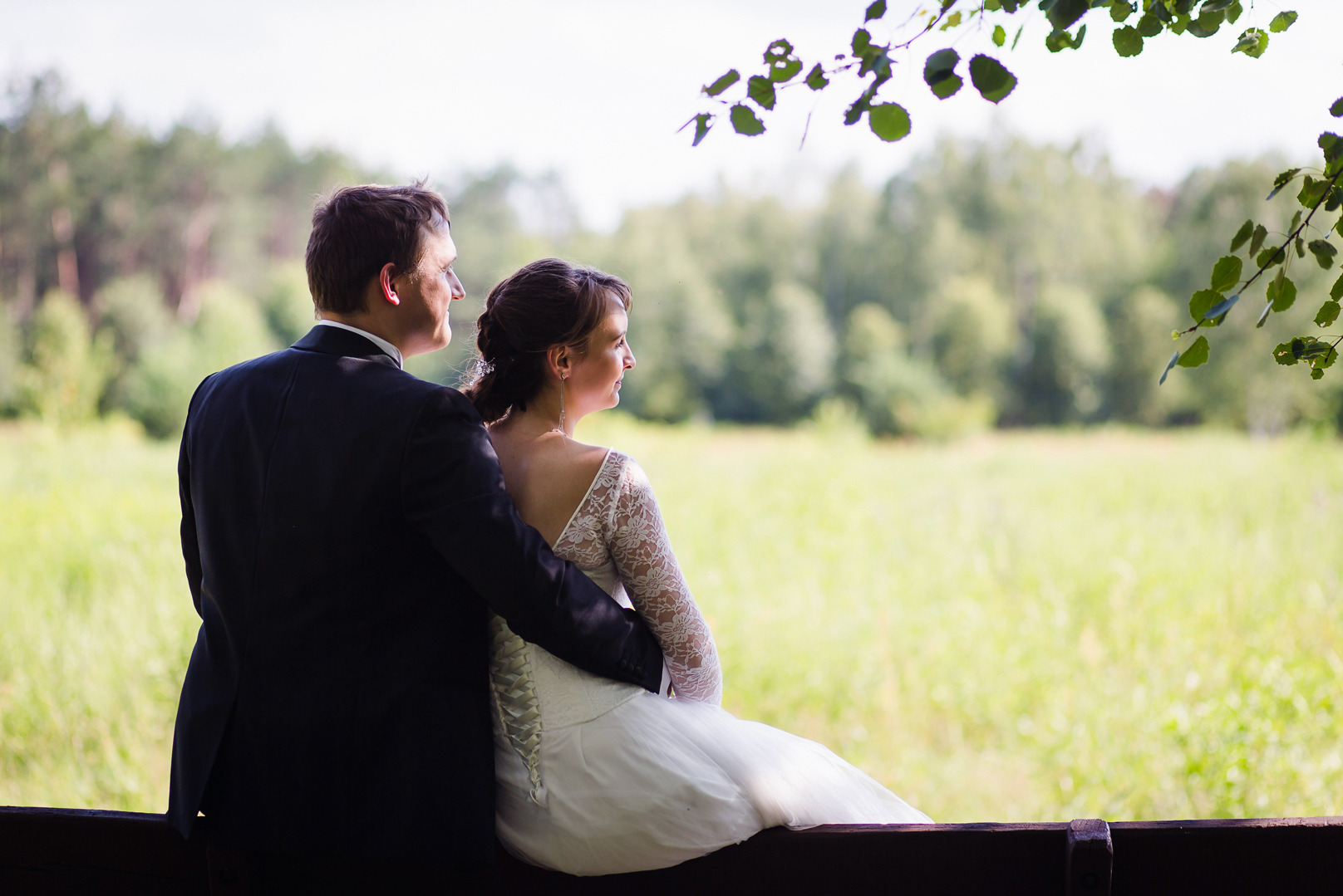 Para młoda w Łodzi siedzi na tle zieleni. Pan w smokingu obejmuje pannę młodą w koronkowej sukni, oboje patrzą w dal. Romantyczna sesja.