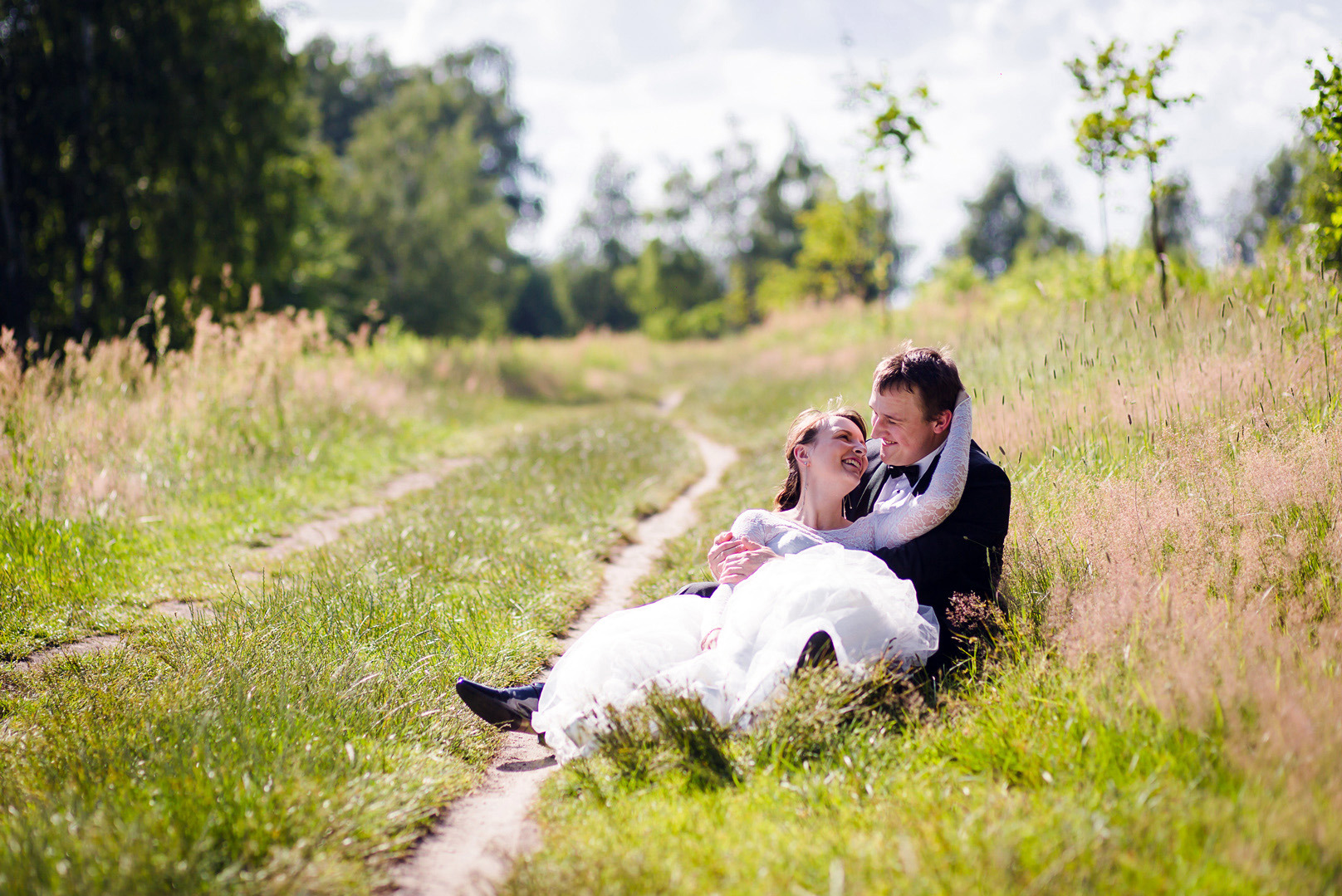 Uśmiechnięta para młoda w objęciach na zielonej łące, on w smokingu, ona w białej sukni, ścieżka w trawie w tle, słoneczny dzień.