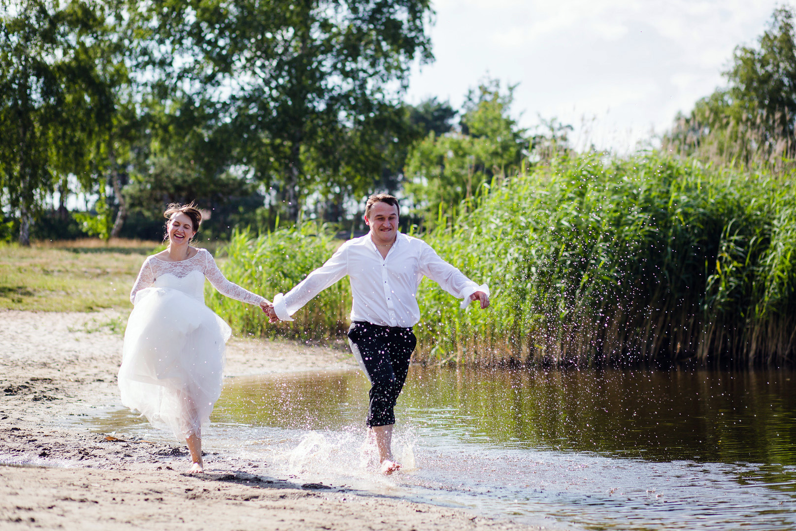 Radosna para młoda biegnie brzegiem jeziora, trzymając się za ręce. Panna młoda w zwiewnej sukni, pan młody w białej koszuli. Woda rozpryskuje się wokół nich.