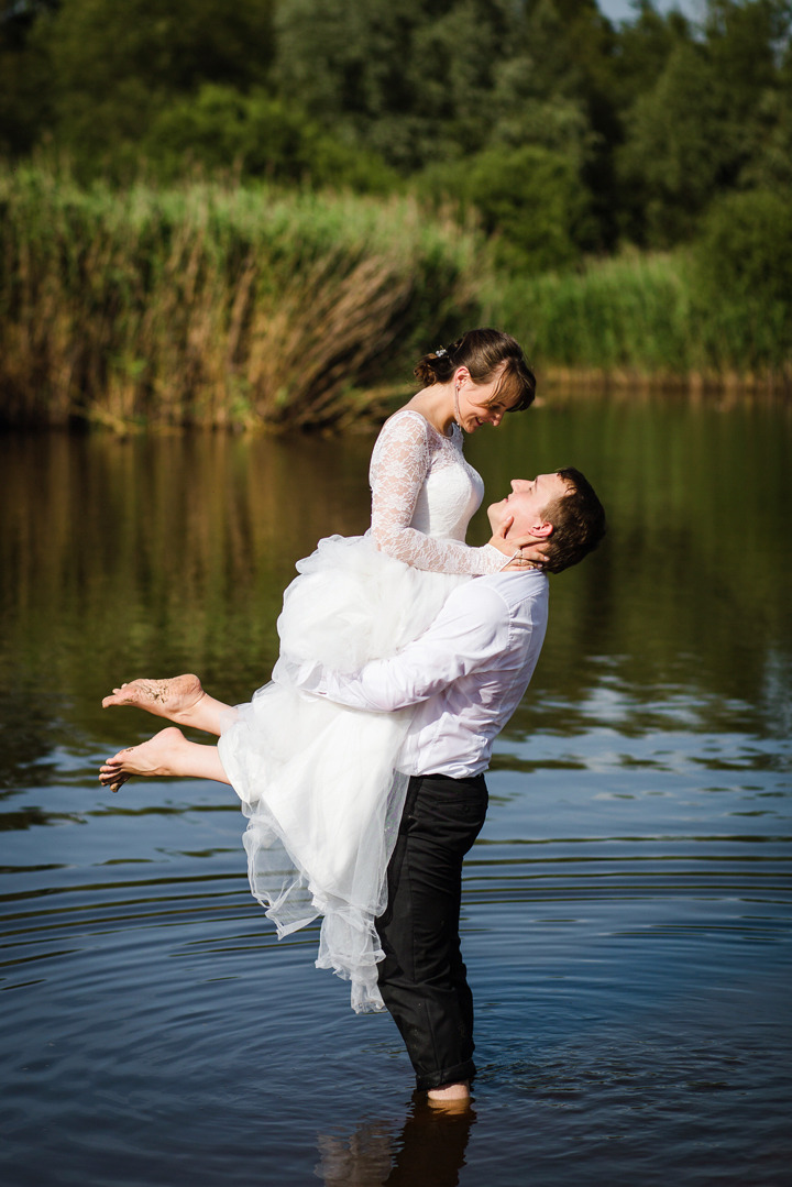 Uśmiechnięty pan młody trzyma pannę młodą w białej sukni na rękach, stojąc w wodzie jeziora na tle trzcin i drzew, w słoneczny dzień. Romantyczna sesja ślubna.