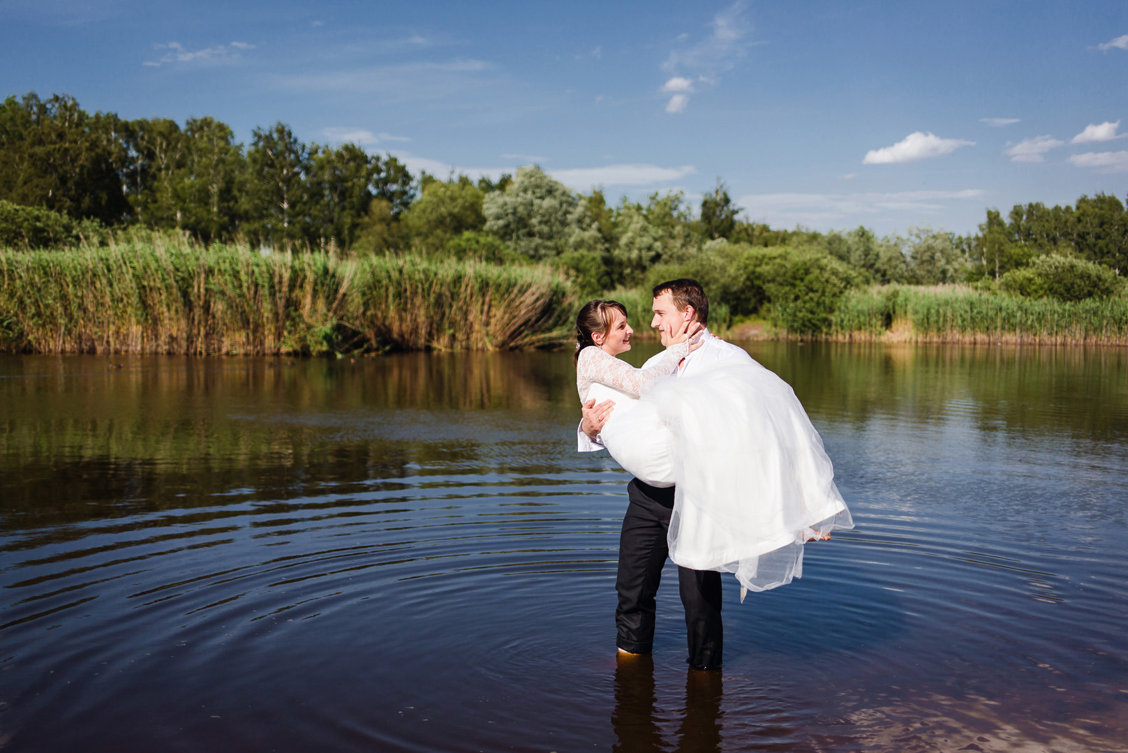 Para młoda w wodzie: pan młody trzyma pannę młodą na rękach, oboje patrzą na siebie z uśmiechem, w tle jezioro i zieleń. Romantyczna sesja ślubna w plenerze.