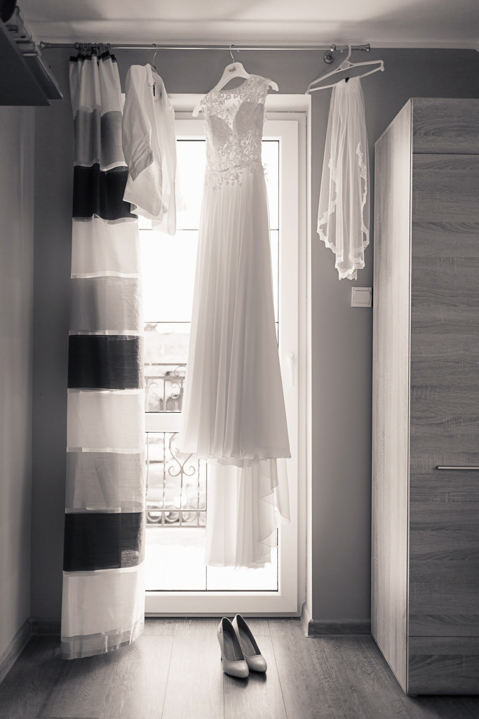 Elegancka suknia ślubna z koronkową górą i welonem wisząca na wieszaku przy oknie, obok biała koszula i buty na podłodze. Stonowana kolorystyka.