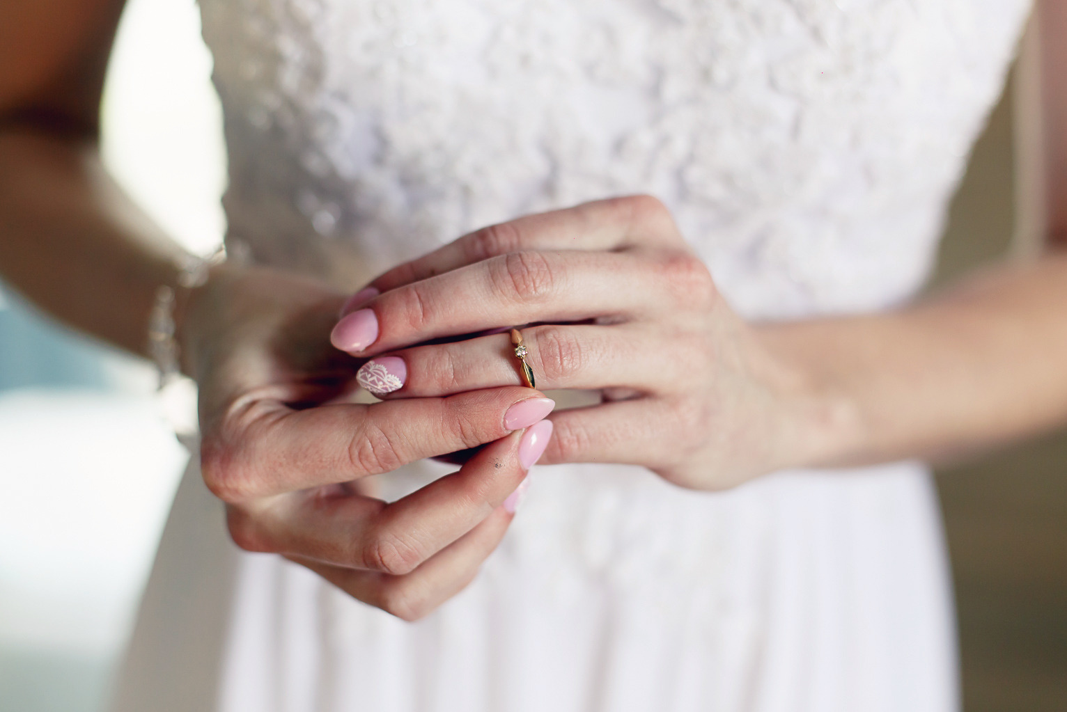 Panna młoda w białej sukni trzyma delikatny złoty pierścionek zaręczynowy, eksponując manicure z koronkowym wzorem na jednym paznokciu.