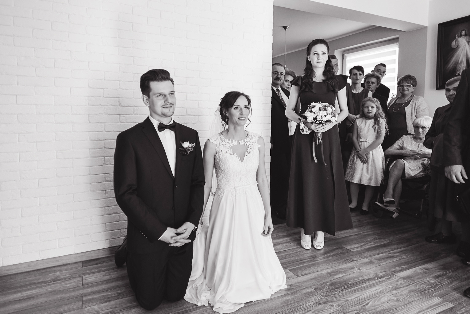 Czarno-białe zdjęcie pary młodej klęczącej na drewnianej podłodze, z gośćmi weselnymi w tle, w jasnym wnętrzu z ceglaną ścianą.