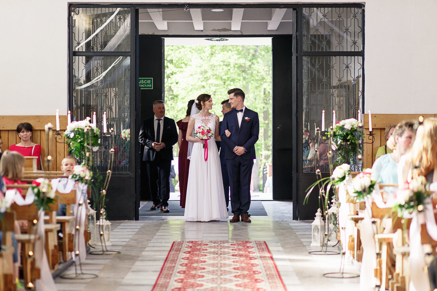 Młoda para w kościele, prowadzona przez mężczyznę w garniturze. Widok na nawę, gości i dekoracje kwiatowe. Jasne, naturalne światło.