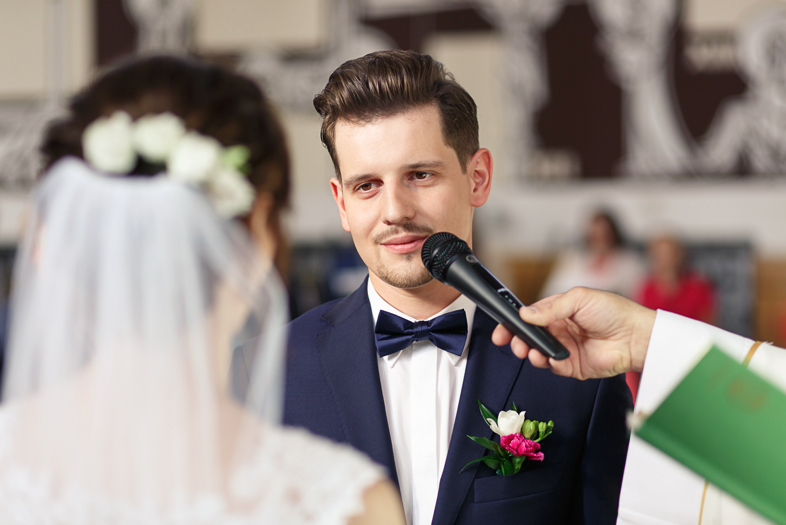 Uśmiechnięty pan młody w granatowym garniturze i muszce, podczas ceremonii ślubnej w kościele. W tle rozmazana sylwetka panny młodej w welonie.