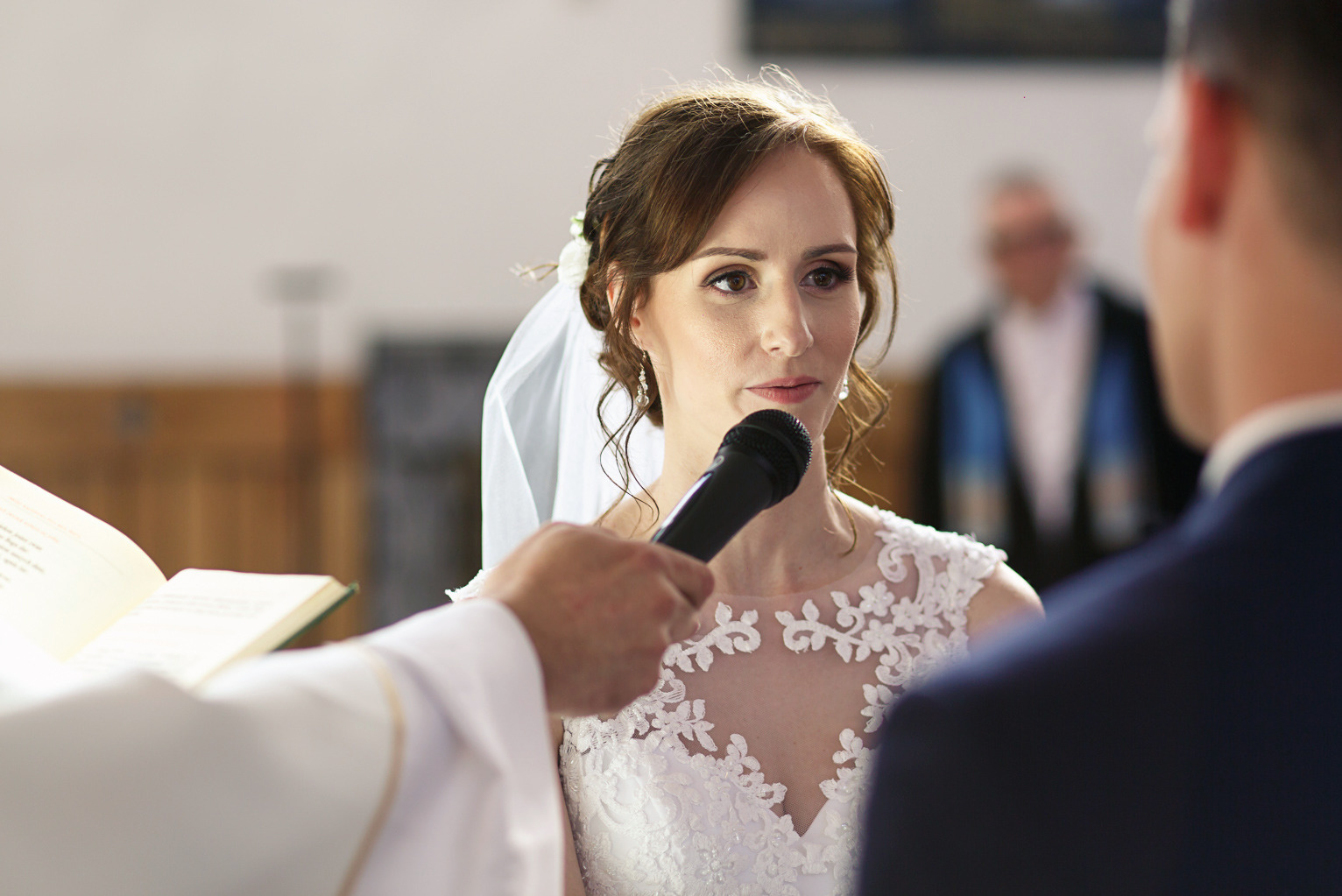 Panna młoda w białej sukni z koronkowym dekoltem, trzyma mikrofon podczas ceremonii ślubnej w kościele. W tle rozmyta postać w stroju liturgicznym.