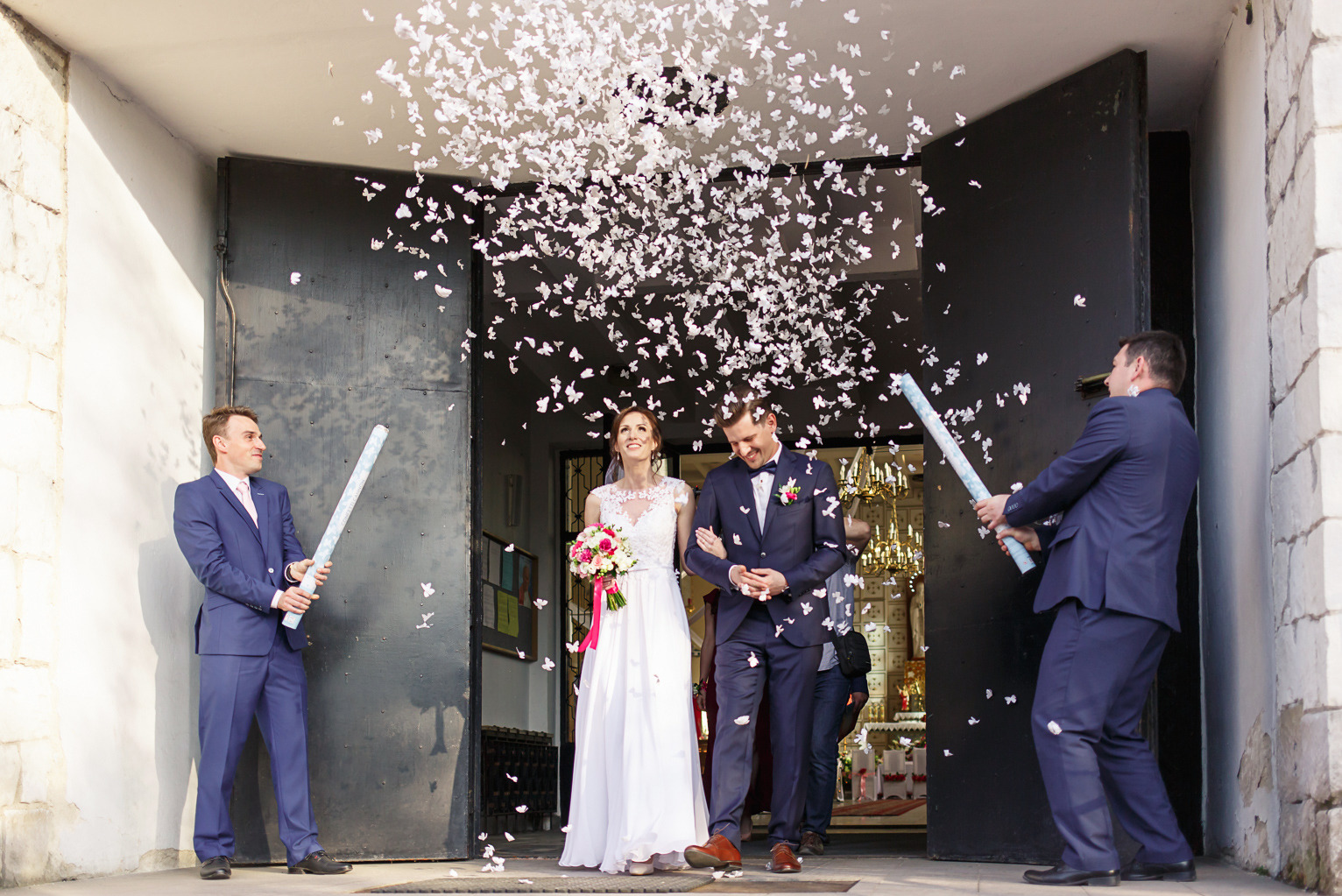 Uśmiechnięta para młoda wychodzi z kościoła obsypywana konfetti w kształcie motyli, w tle widoczni mężczyźni w garniturach z tubami konfetti.