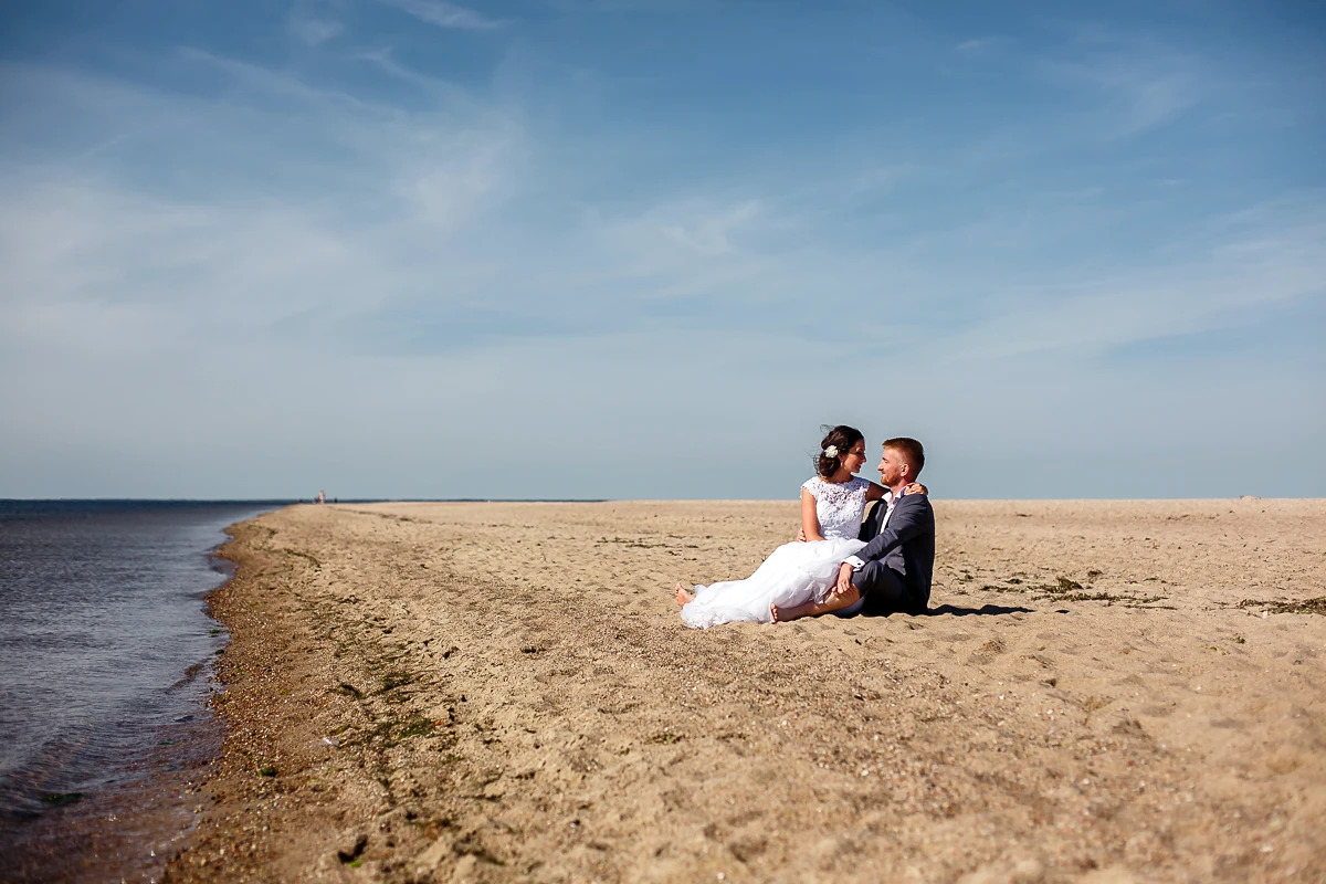 Para młoda siedzi na piaszczystej plaży nad brzegiem morza w słoneczny dzień. Panna młoda w białej sukni, pan młody w szarym garniturze, oboje patrzą na siebie z uśmiechem.
