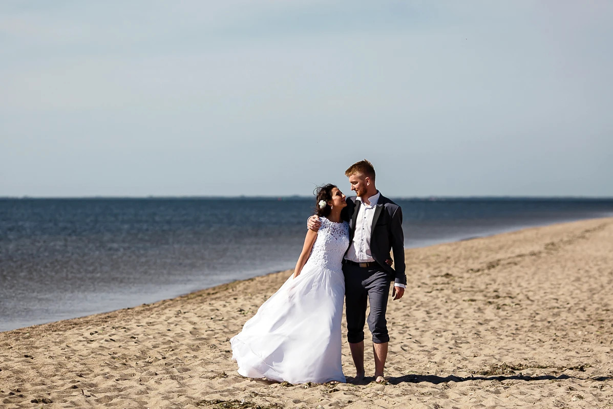 Para młoda na plaży: Panna młoda w białej sukni z koronkową górą, Pan młody w szarym garniturze bez skarpet, patrzą na siebie z czułością na tle morza.