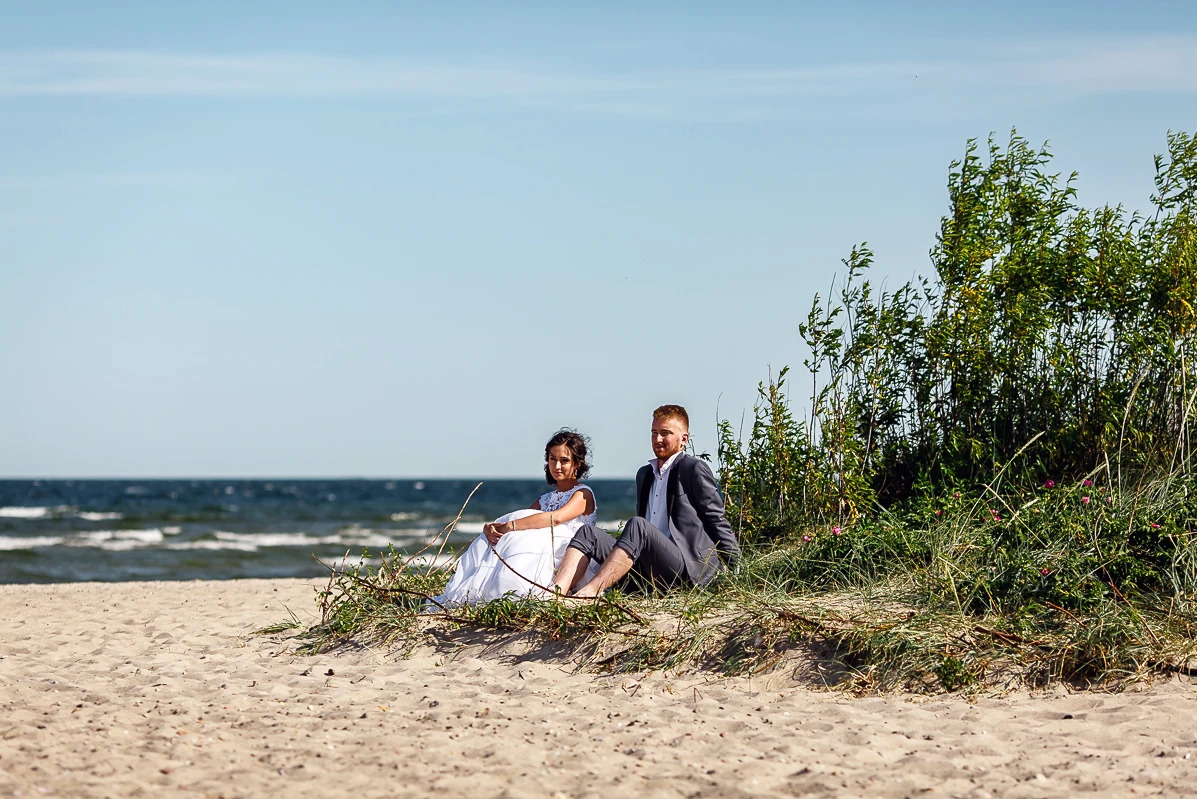 Para młoda siedzi na piaszczystej plaży z widokiem na morze. Kobieta w białej sukni, mężczyzna w szarym garniturze. Romantyczna sesja na tle błękitnego nieba.