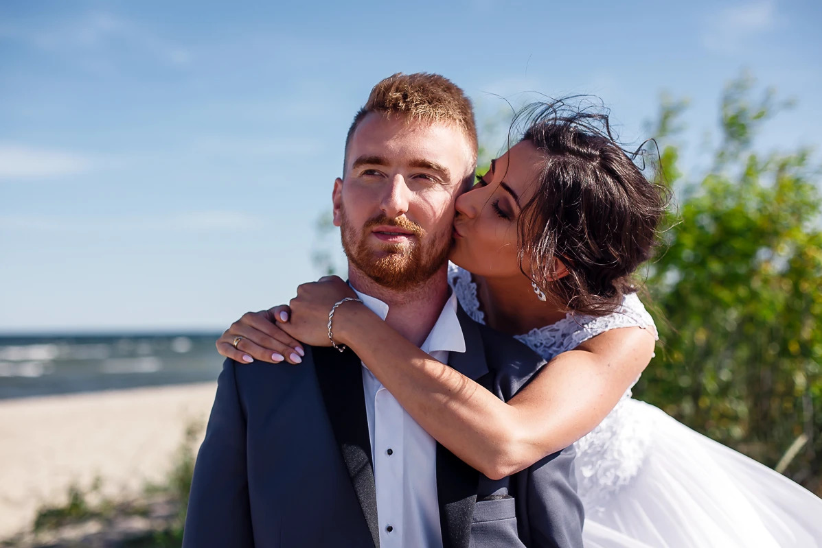 Panna Młoda w koronkowej sukni całuje w policzek Pana Młodego w ciemnym garniturze na tle plaży i morza. Ujęcie z sesji plenerowej.