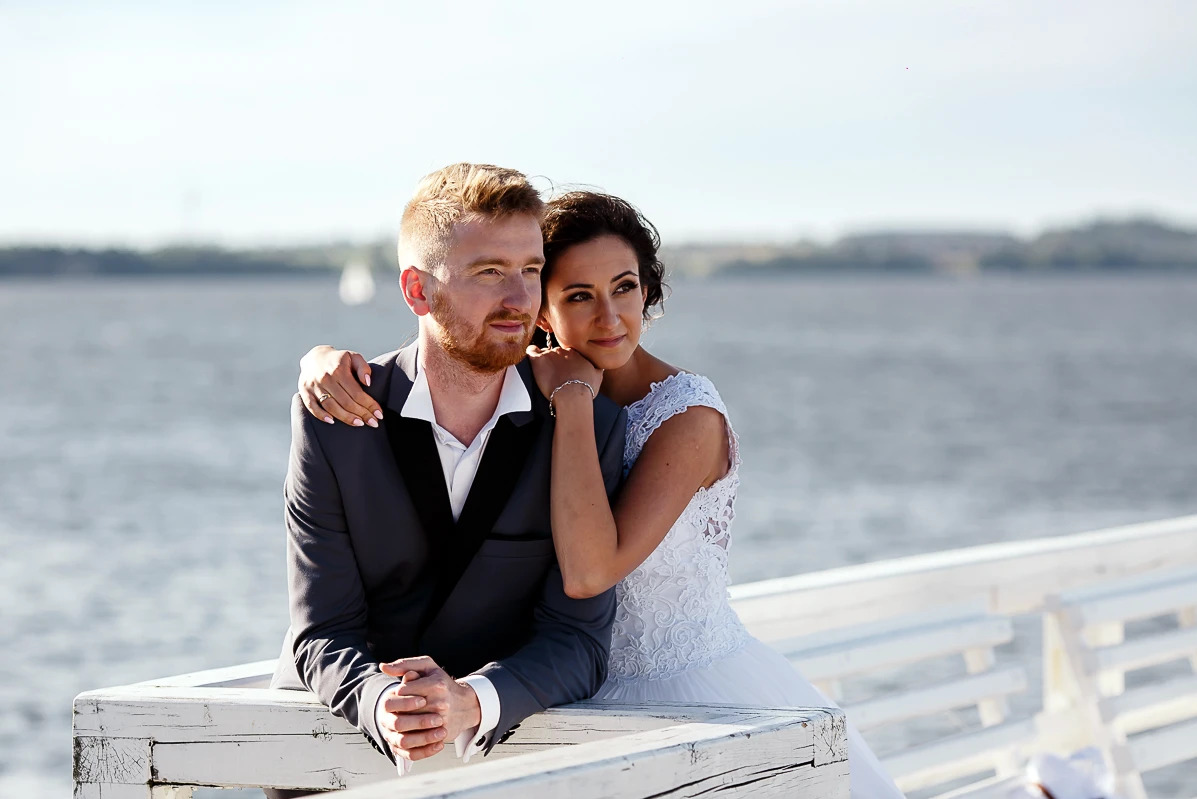 Para młoda w eleganckich strojach pozuje na molo z widokiem na spokojne morze w słoneczny dzień. Delikatne oświetlenie podkreśla detale sukni.