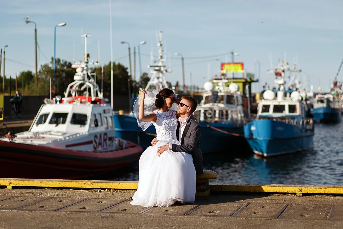 Para młoda w objęciach na tle portu z łodziami SAR, w słoneczny dzień. Panna młoda w koronkowej sukni i welonie, pan młody w garniturze i okularach.