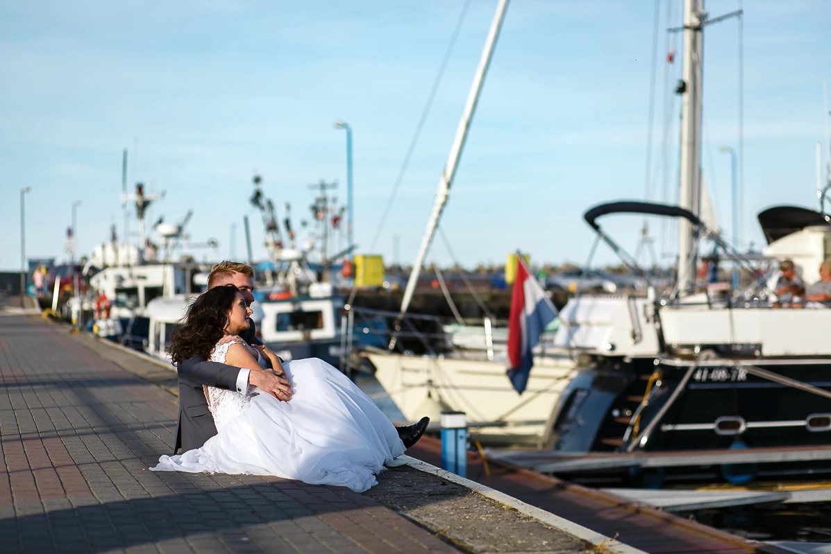 Para młoda siedzi na nabrzeżu portowym, mężczyzna trzyma kobietę w sukni ślubnej na rękach, w tle jachty i błękitne niebo.