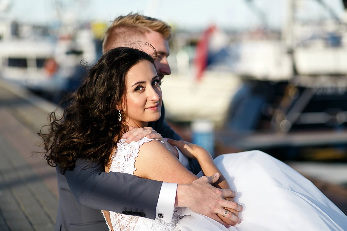 Uśmiechnięta panna młoda w objęciach pana młodego na tle portu jachtowego. Suknia z koronką, elegancki garnitur, delikatny makijaż, pierścionek na palcu pana młodego.