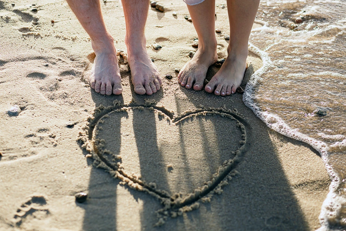 Stopy pary na piasku obok serca narysowanego na plaży, z falami delikatnie obmywającymi brzeg. Romantyczna sesja na tle morza.
