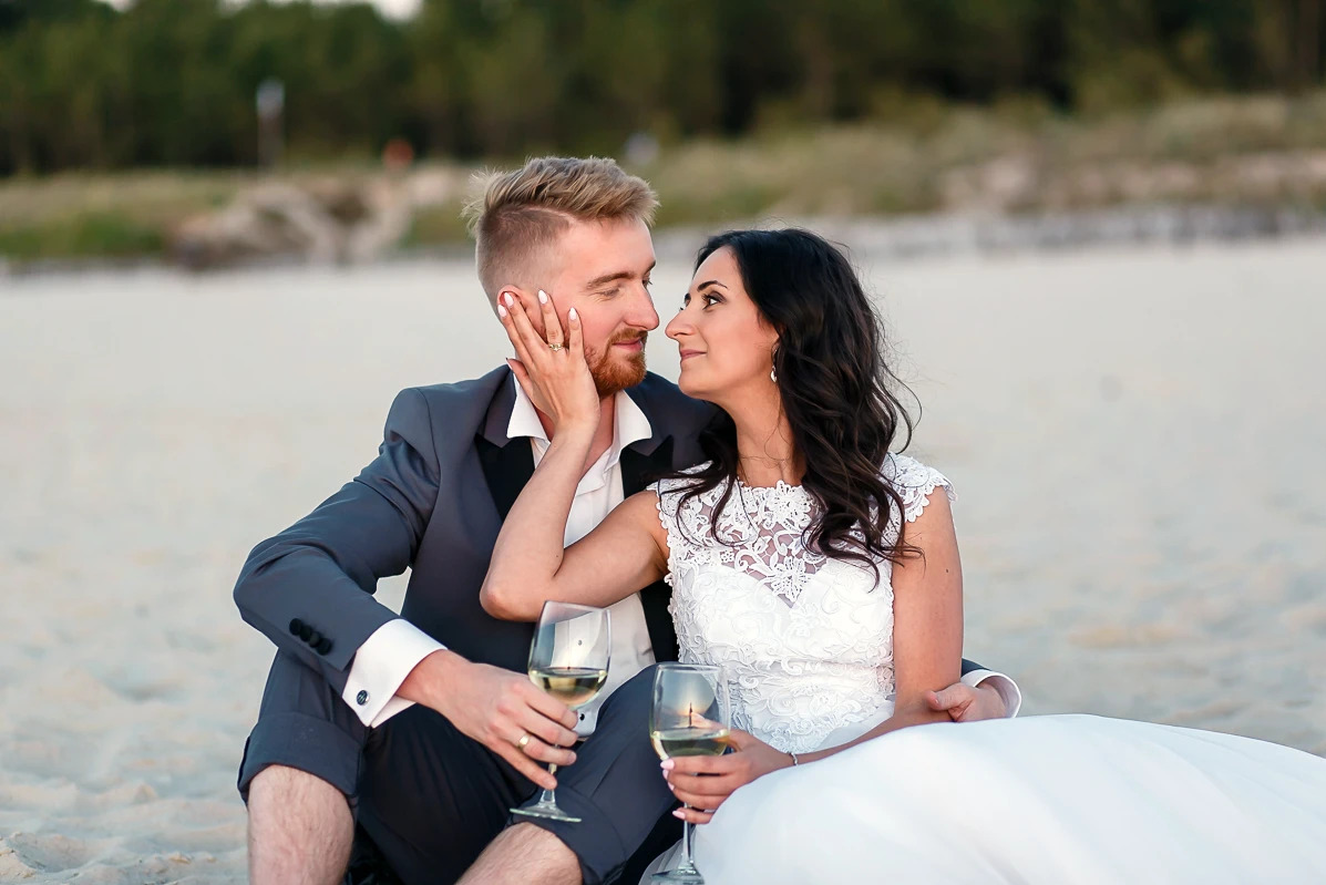 Para młoda siedzi na plaży z kieliszkami wina, patrząc na siebie z czułością. Ona w koronkowej sukni, on w garniturze z podwiniętymi nogawkami, boso. Romantyczna sesja ślubna.