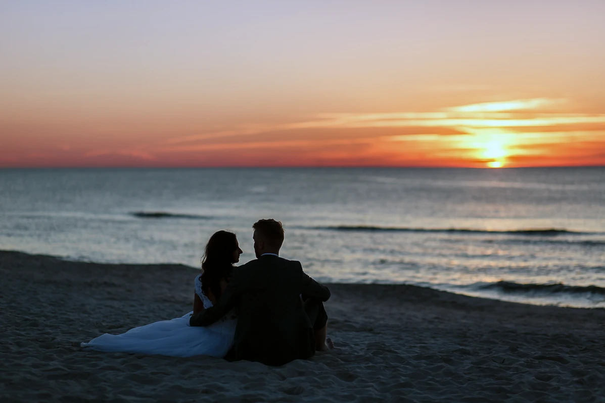 Para młoda siedzi na plaży, obejmując się, na tle zachodzącego słońca nad morzem. Romantyczny kadr z sesji ślubnej w ciepłych barwach.