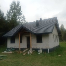 Dachy Gromnicki - Dom w trakcie budowy z ciemną dachówką i drewnianą konstrukcją ganku. Widoczne bloczki betonowe i fragment lasu w tle.