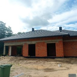 Dachy Gromnicki - Dom w trakcie budowy z ciemną dachówką i widocznymi rynnami, ceglana konstrukcja ścian, teren budowy z piaskiem i zielonym koszem na śmieci.