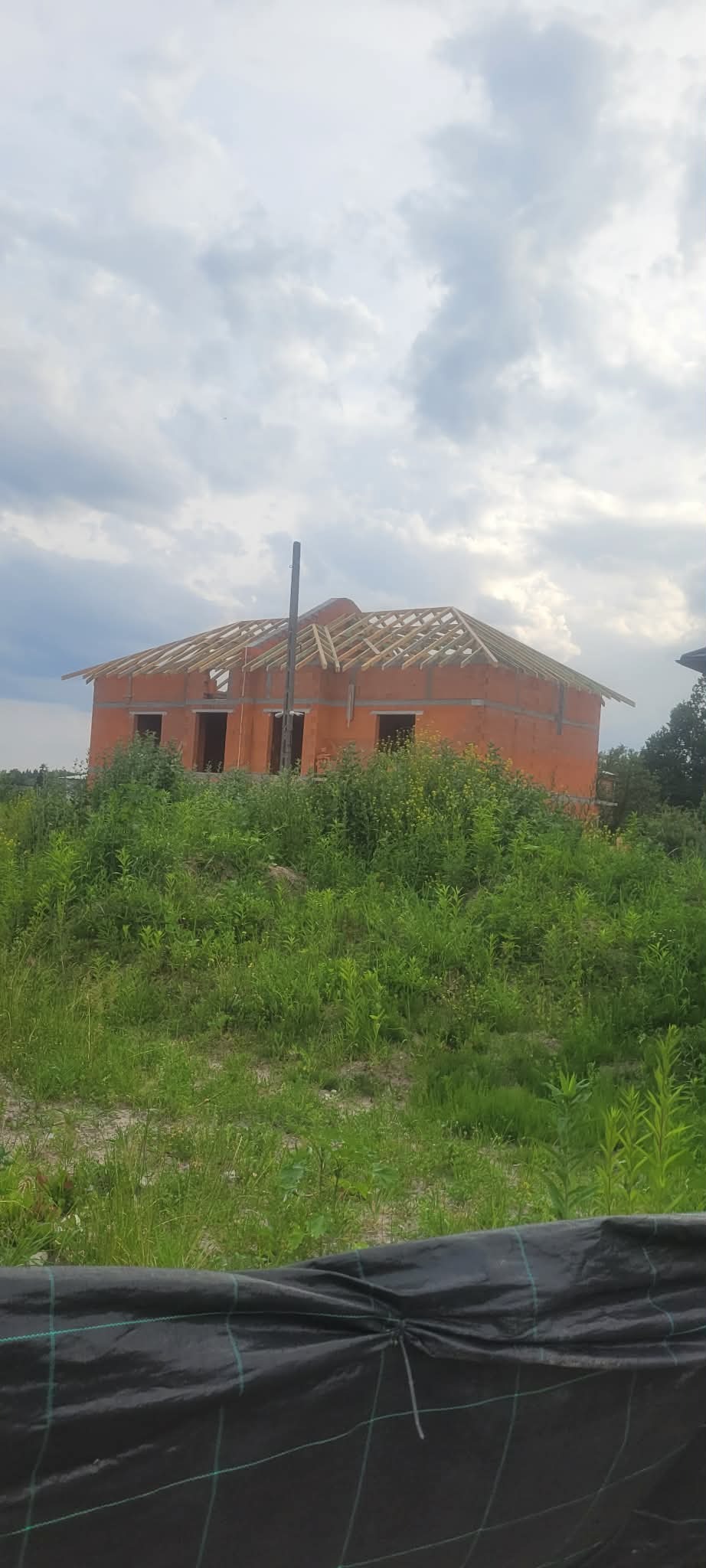 Surowy budynek z cegły z drewnianą konstrukcją dachu w trakcie budowy, otoczony zielenią i pochmurnym niebem. Widoczny komin i fragment czarnej plandeki.