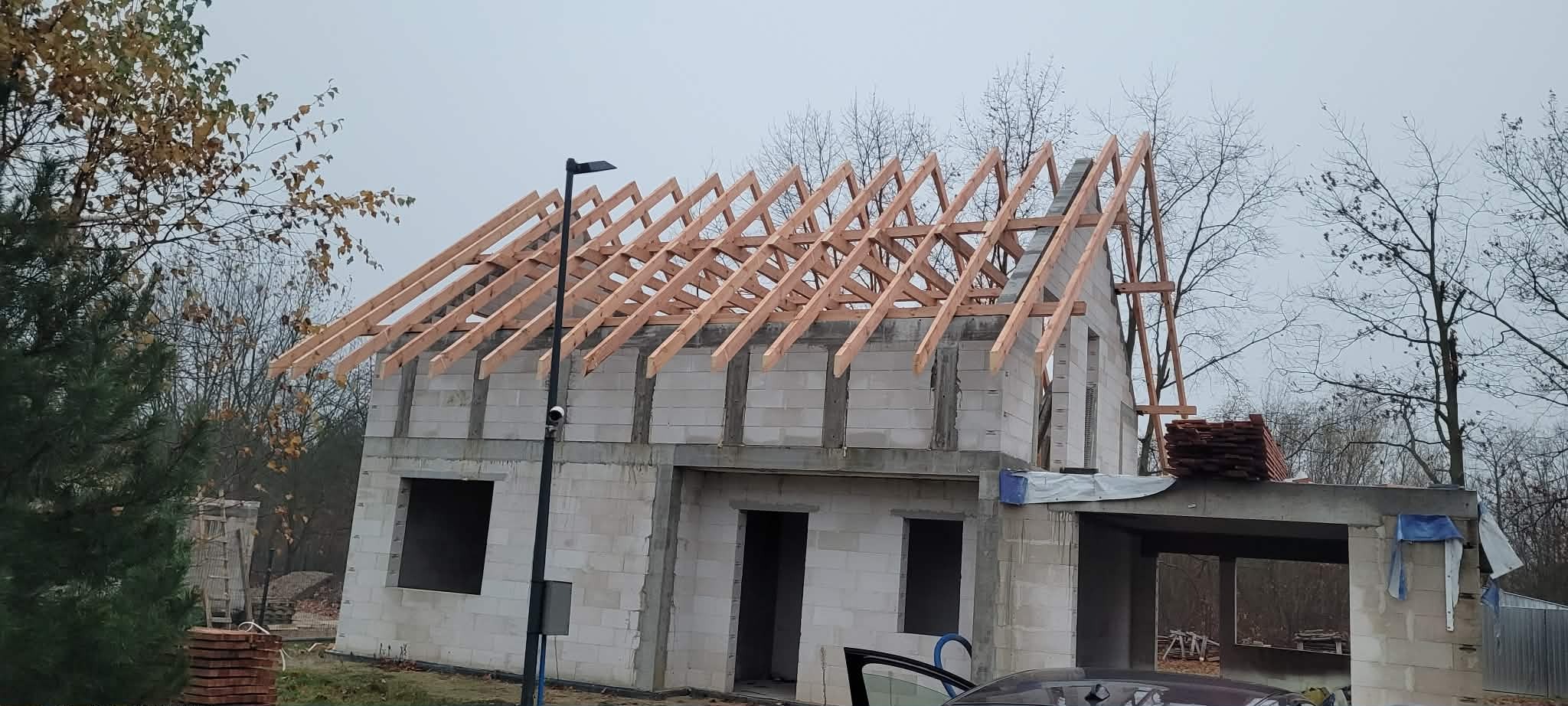 Surowy stan domu jednorodzinnego z widoczną konstrukcją drewnianą dachu w trakcie budowy, z elementami deskowania i przygotowanymi dachówkami. Pochmurny dzień.
