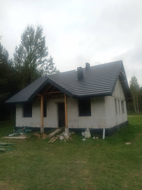 Dom w trakcie budowy z ciemną dachówką i drewnianą konstrukcją ganku. Widoczne bloczki betonowe i fragment lasu w tle.