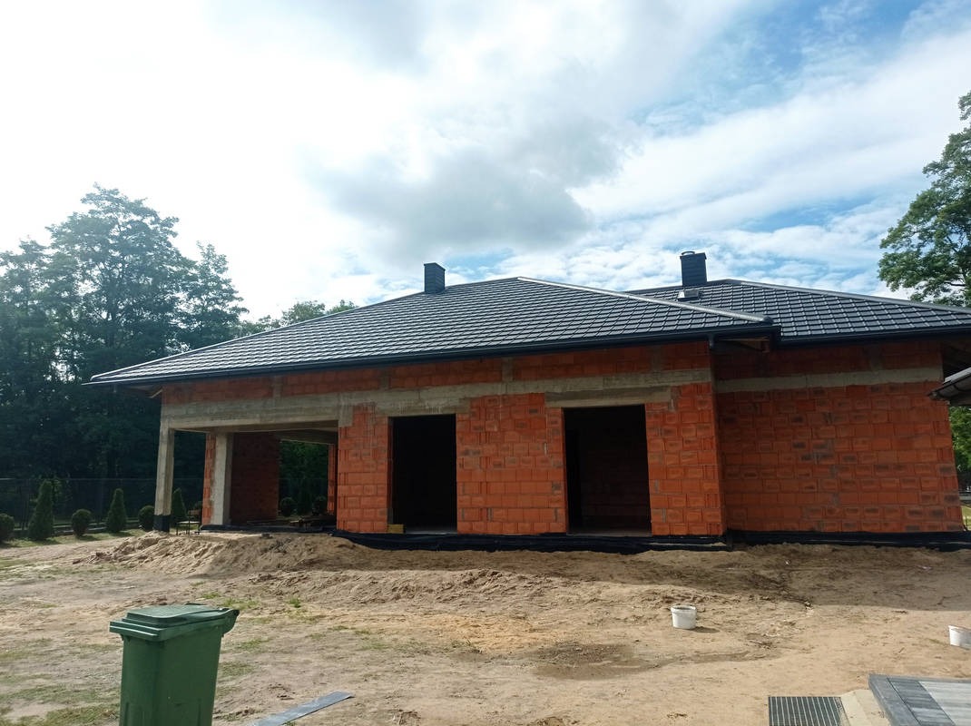 Dom w trakcie budowy z ciemną dachówką i widocznymi rynnami, ceglana konstrukcja ścian, teren budowy z piaskiem i zielonym koszem na śmieci.