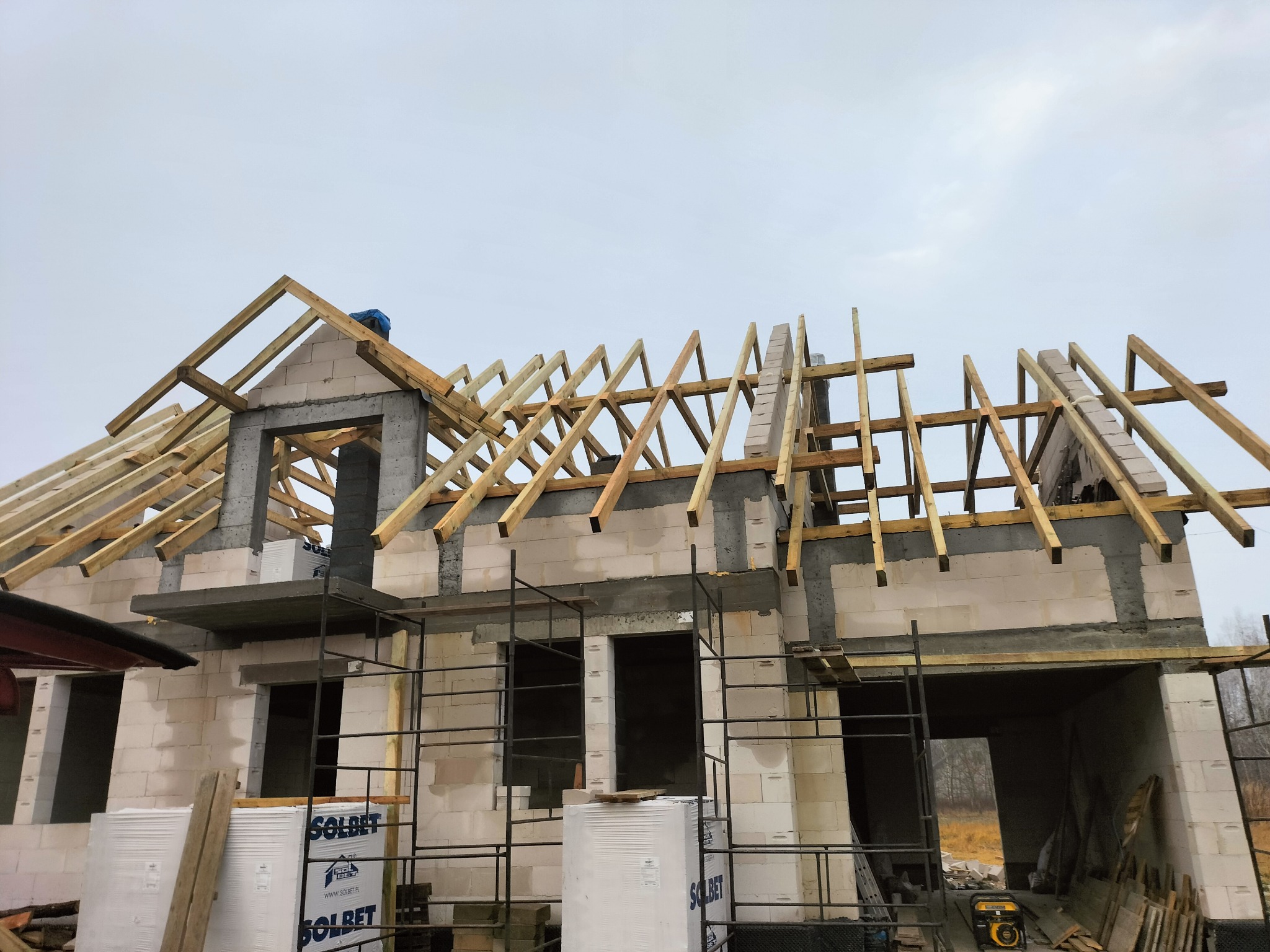 Budowa domu jednorodzinnego: wznoszenie konstrukcji dachu z drewnianych krokwi na ścianach z betonu komórkowego. Widoczne rusztowania i materiały budowlane.