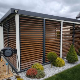 CARGO CENTER SPÓŁKA Z OGRANICZONĄ ODPOWIEDZIALNOŚCIĄ - Nowoczesna, zamknięta pergola z drewnianymi żaluzjami, otoczona dekoracyjnymi krzewami i białym żwirem. Widoczny zwijany wąż ogrodowy i kamera na elewacji.