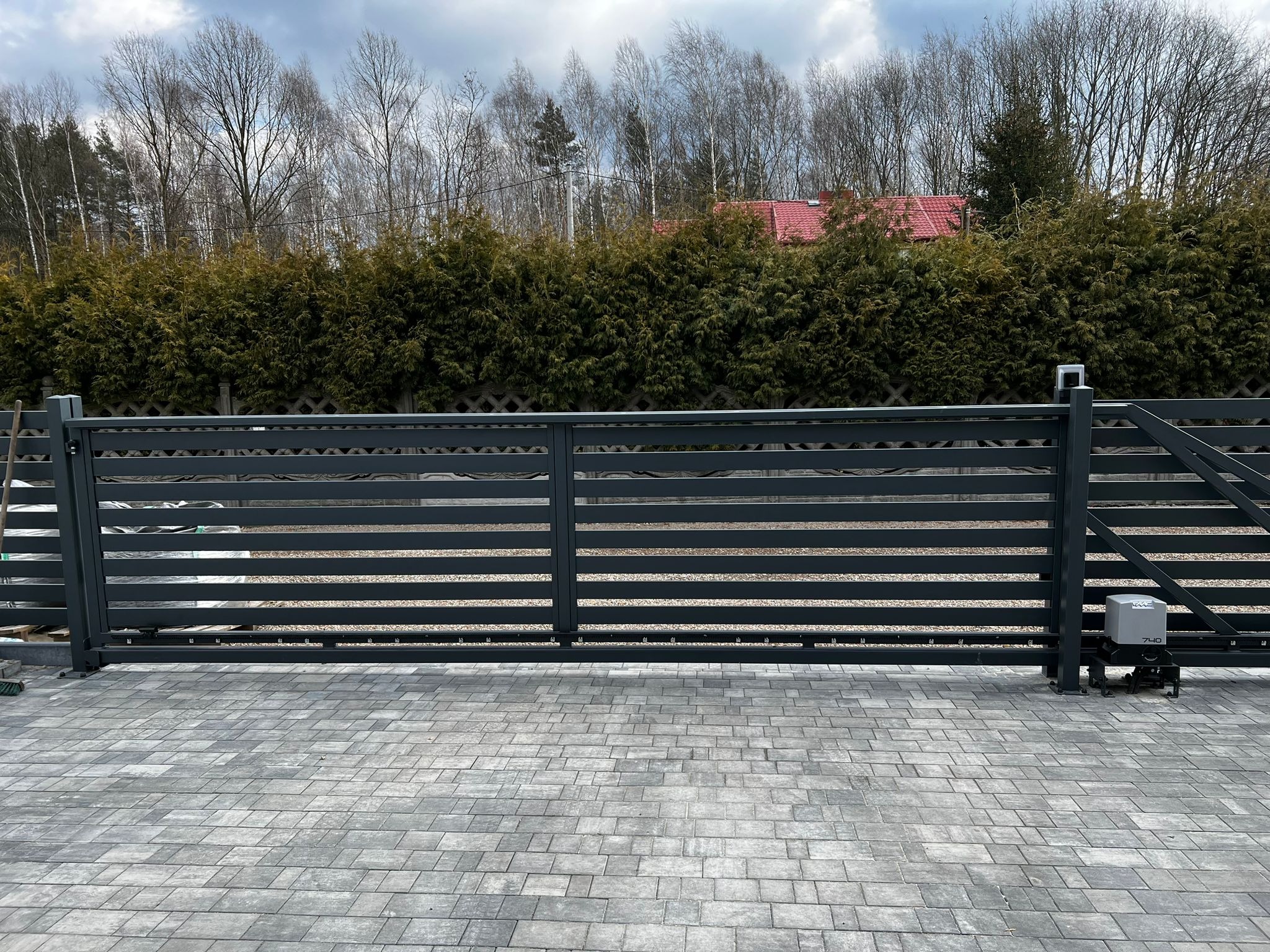 Nowoczesna, przesuwna brama wjazdowa z automatyką, szara, poziome profile. Widoczny napęd bramy. Tło: zielony żywopłot i drzewa. Kostka brukowa.