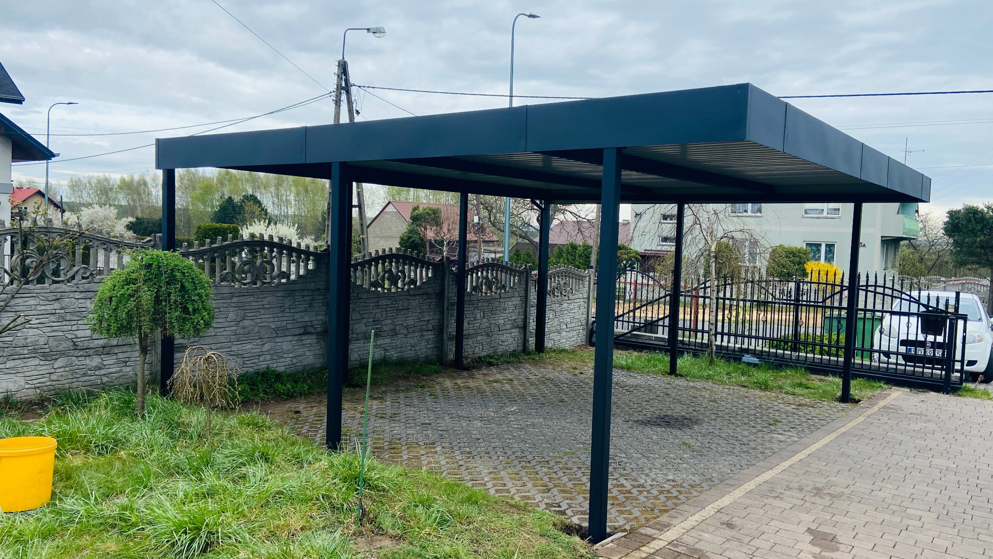 Nowoczesna, grafitowa wiata garażowa z metalową konstrukcją na posesji z kostki brukowej, otoczona zielenią i ogrodzeniem z kamienia. W tle domy.