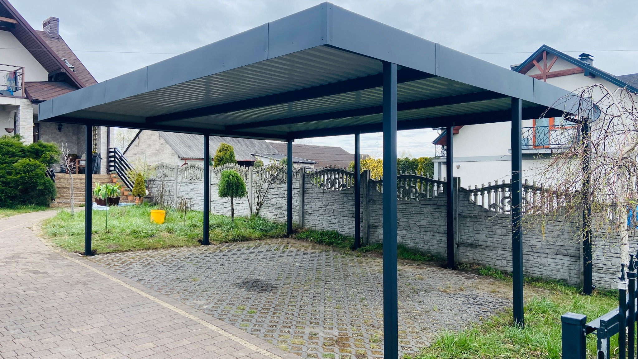 Nowoczesna, grafitowa konstrukcja z metalu, pełniąca funkcję zadaszenia nad podjazdem, z widocznym ażurowym brukiem i fragmentem ogrodzenia.